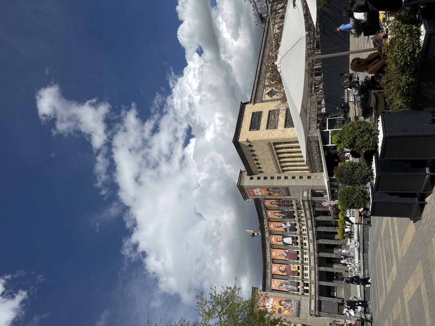 This daytime image captures a vibrant urban scene in Yerevan, Armenia, featuring the Moscow Cinema, an outdoor cafe, and a clear sky with prominent clouds.
The upper portion of the frame is dominated by a deep blue sky, accented by large, voluminous white cumulus clouds. Below the sky, the Moscow Cinema building forms a significant part of the mid-ground. It showcases a grand architectural style with a curved facade featuring a colonnade. Numerous large promotional banners for films or events adorn the facade, displaying text in both Cyrillic and Armenian scripts. Clearly visible Cyrillic text spells out "МОСКВА" (Moscow) and "КИНОТЕАТР" (Cinema) on one of the central banners, confirming the building's identity. Above this curved section, the building continues with more rectangular sections, detailed with windows and decorative elements.
To the right side, an active outdoor cafe or seating area is visible. This area is furnished with white patio umbrellas providing shade over tables and chairs. Planters filled with lush green bushes and small trees delineate the space. The ground is paved with light-colored square tiles.
Several people are present in the scene. Some are seated at the cafe, enjoying the outdoor setting, while others are walking on the paved area in front of the cinema and adjacent to the cafe. Their attire suggests casual wear suitable for pleasant weather.
The bright sunlight indicates the time of day is mid-morning to early afternoon, casting clear shadows and illuminating the scene. The overall atmosphere is lively and inviting, typical of a bustling city center enjoying good weather.