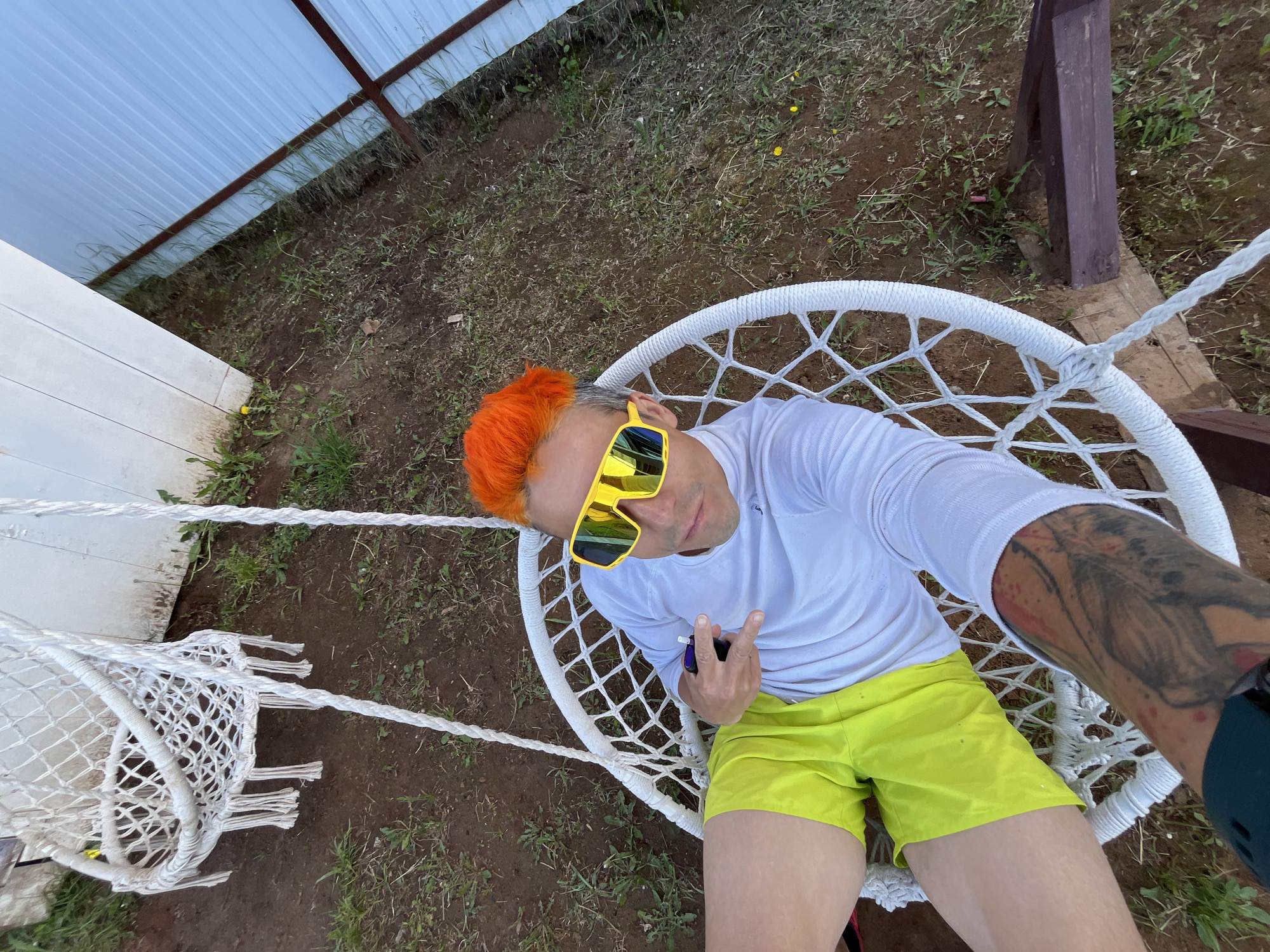 A person is photographed from above while seated in a white rope swing chair, possibly located in Chaikovsky, Russia, given the user's location. The person has bright orange hair, yellow sunglasses, a white long-sleeve shirt, and bright yellow shorts. They are holding a dark-colored object in their left hand, making a gesture with their fingers. Their right arm is extended towards the camera, revealing a tattoo. The swing is suspended from a wooden structure and white rope. The backdrop consists of a yard with a metal fence and some greenery. The ground appears to be a mix of dirt and grass.