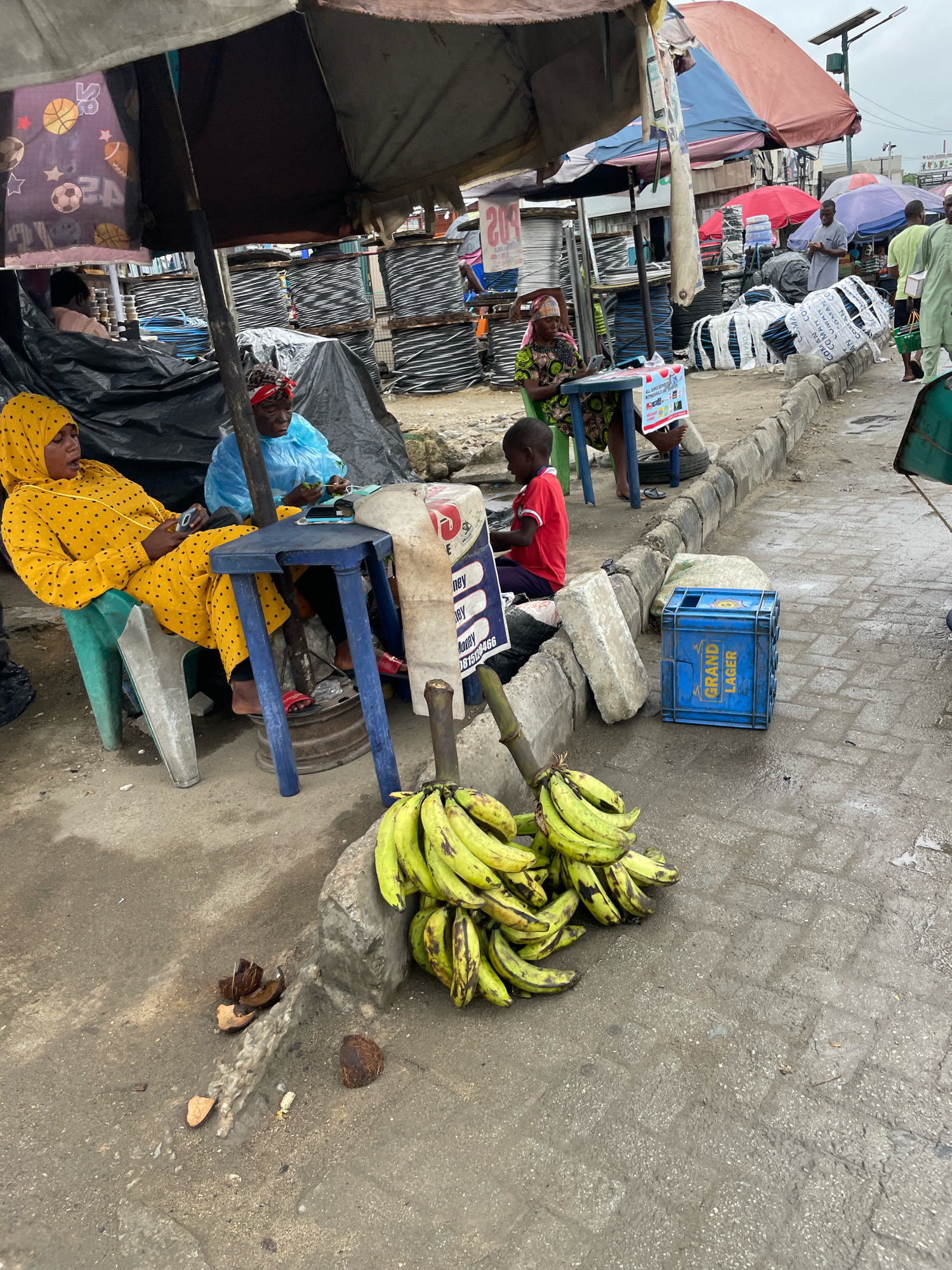 In Ajah, Nigeria, a street market scene unfolds under makeshift awnings. Two women are seated, one in a yellow patterned headscarf and dress, holding a mobile phone, and another in a blue plastic covering, appearing to be engaged in conversation or a transaction. A young boy in a red shirt sits near them, facing away from the camera. In front of them, a small blue table holds a mobile phone and other items. Beside the table, a large white bag with blue and red lettering is visible.
On the ground, a pile of green bananas rests on a concrete curb. Nearby, a blue plastic crate labeled "GRAND LAGER" sits on the wet pavement. Several large spools of what appear to be wire or cable are stacked behind the people, covered by black tarpaulin. Scattered debris, including pieces of coconut shells, are also present on the ground. The environment suggests a busy, outdoor marketplace with various goods and a somewhat overcast sky.