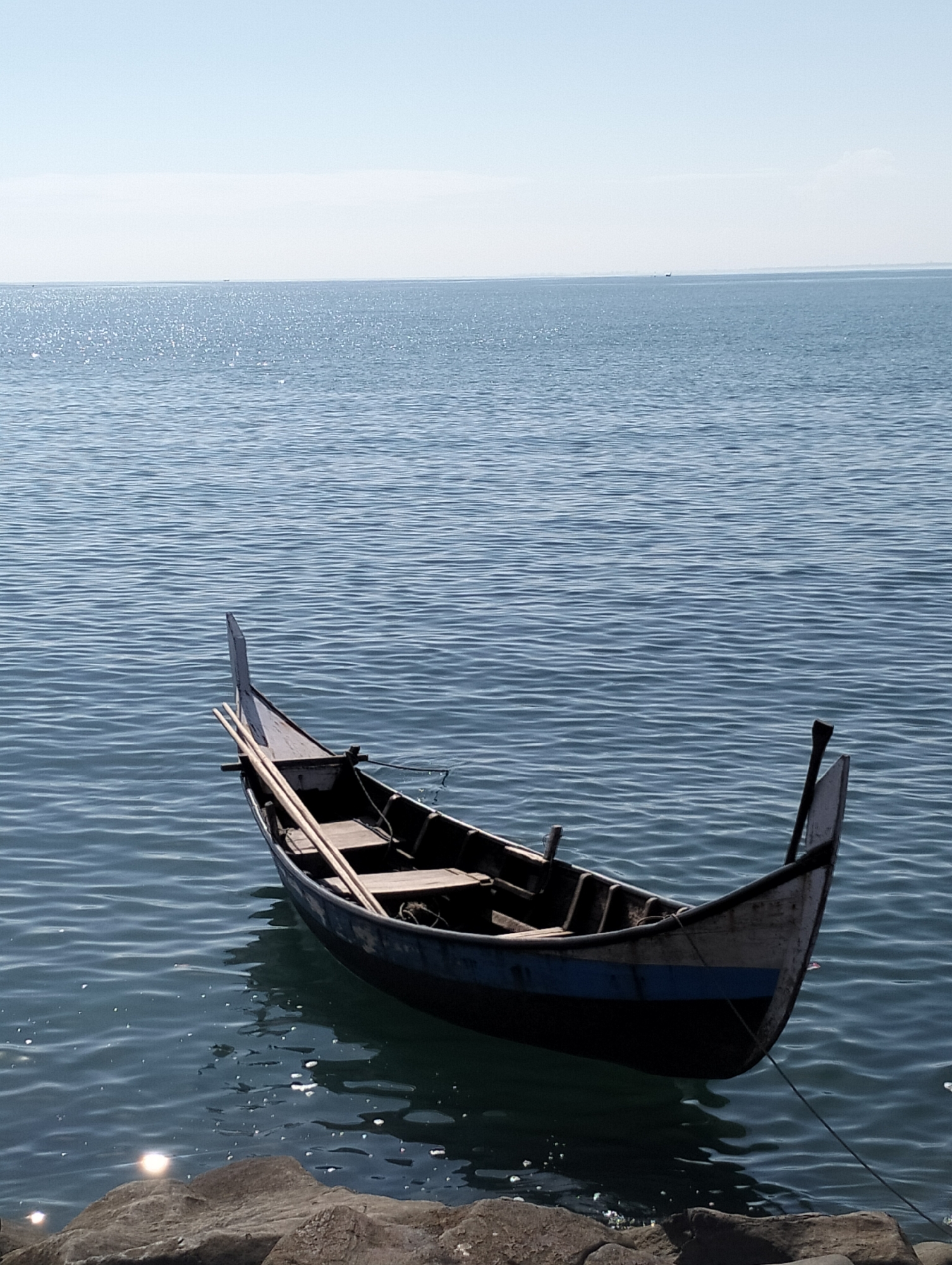 A slightly high-angle shot depicts a small boat afloat on the sea, near the city of Lhokseumawe, Indonesia. The boat, painted predominantly black with a white trim and a horizontal blue stripe, has two poles resting inside. It is situated close to a rocky shoreline in the foreground. The sea behind the boat is marked by small waves and the sky is clear and light blue. The sun's reflection can be seen in the water near the rocks.