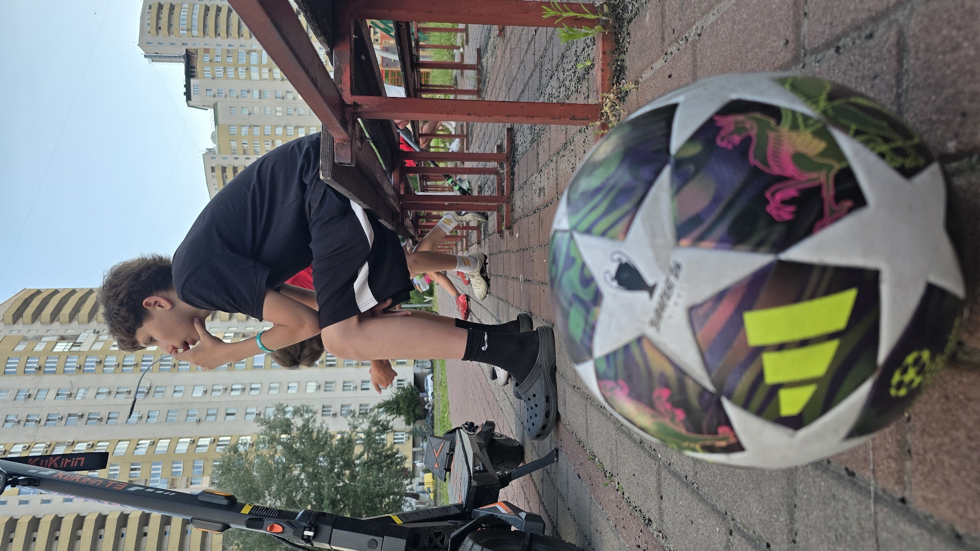 A young boy, wearing a black t-shirt and shorts, leans over a red wooden bench outdoors. He is holding his hand to his chin, appearing thoughtful or perhaps tired. A soccer ball with a vibrant pattern of white stars and colorful designs, including pink figures and yellow stripes, rests on the ground next to him. In the background, tall, light-colored apartment buildings with many windows dominate the skyline. The scene appears to be in a residential area. A scooter is visible in the foreground, partially obscuring the boy's legs. The lighting suggests it is daytime, possibly late afternoon or early evening, with a bright, overcast sky. The ground is paved with brick. The image is taken in Kyiv, Ukraine. Some text is visible on the scooter, reading "Kukirin T3".