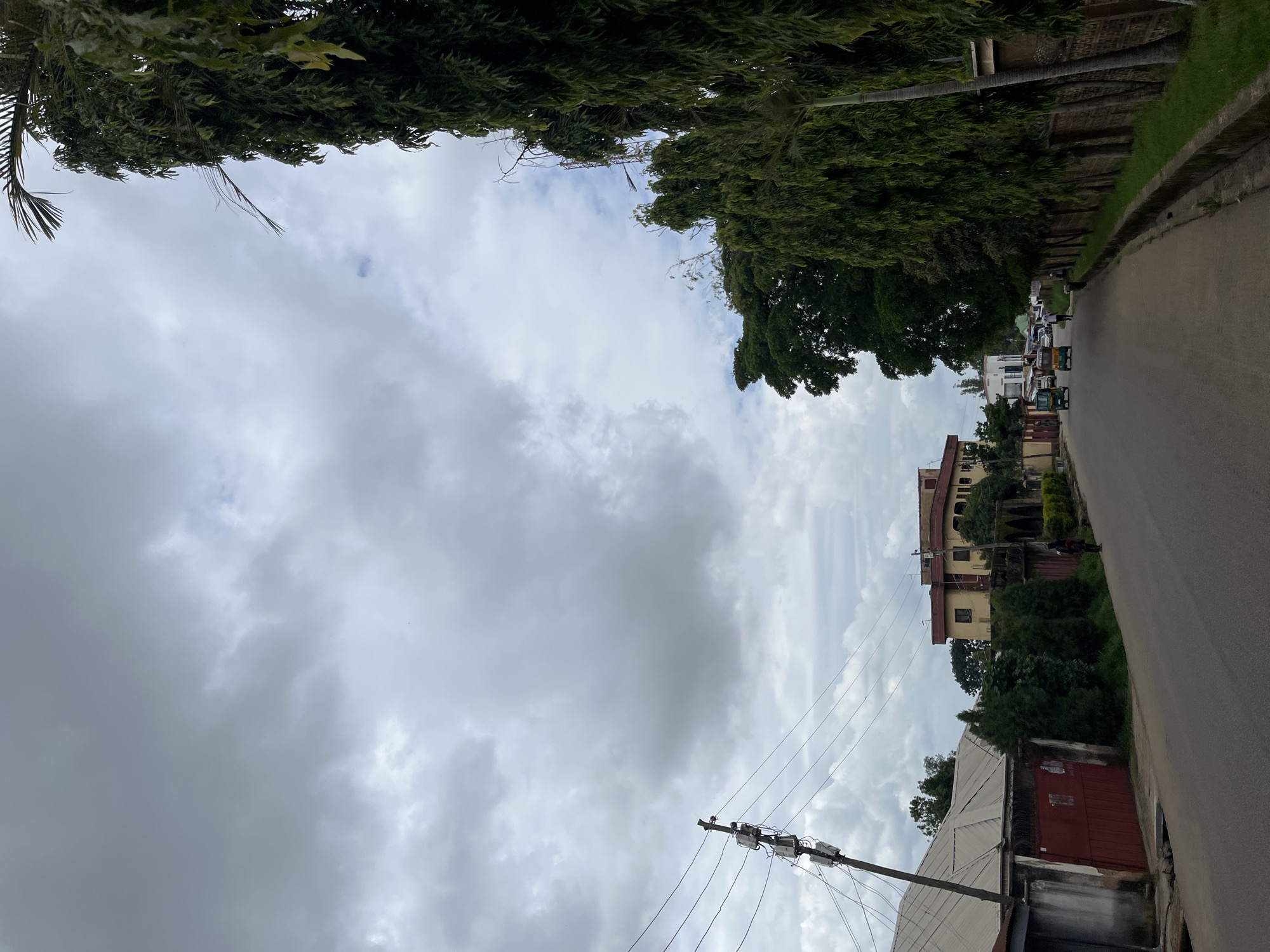 A street scene in Jos, Nigeria, is depicted under a cloudy sky. The perspective is from a low angle, looking upwards. The left side of the image is dominated by lush green trees, their branches forming a dense canopy. On the right side, a paved road runs alongside a line of buildings and more trees. Several vehicles, including what appear to be auto-rickshaws, are visible on the road, suggesting moderate traffic. The buildings are varied in style and color, with some appearing to be multi-story residential structures. Overhead, a telephone pole with wires and electrical boxes is prominent against the cloudy sky. The overall impression is of a typical urban street in a tropical or subtropical region. The weather appears overcast, with diffused light, indicating it might be morning or late afternoon. No people are clearly discernible in detail, though figures are suggested in some of the vehicles and near the buildings. The scene is calm and ordinary, capturing a moment of everyday life in the city.