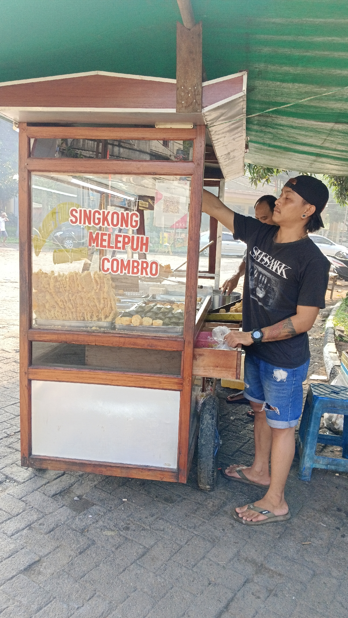 In Tajurhalang, Indonesia, a food cart vendor stands beside a wooden and glass display case filled with fried snacks, including what appears to be "Singkong Melepeh Combo" as indicated by text on the glass. The vendor, wearing a black t-shirt, denim shorts, and a black cap, is reaching into the cart. Behind him, another person is visible, also attending to the food. The cart has a green canopy and is parked on a paved surface. The environment suggests an outdoor market or roadside setting.
