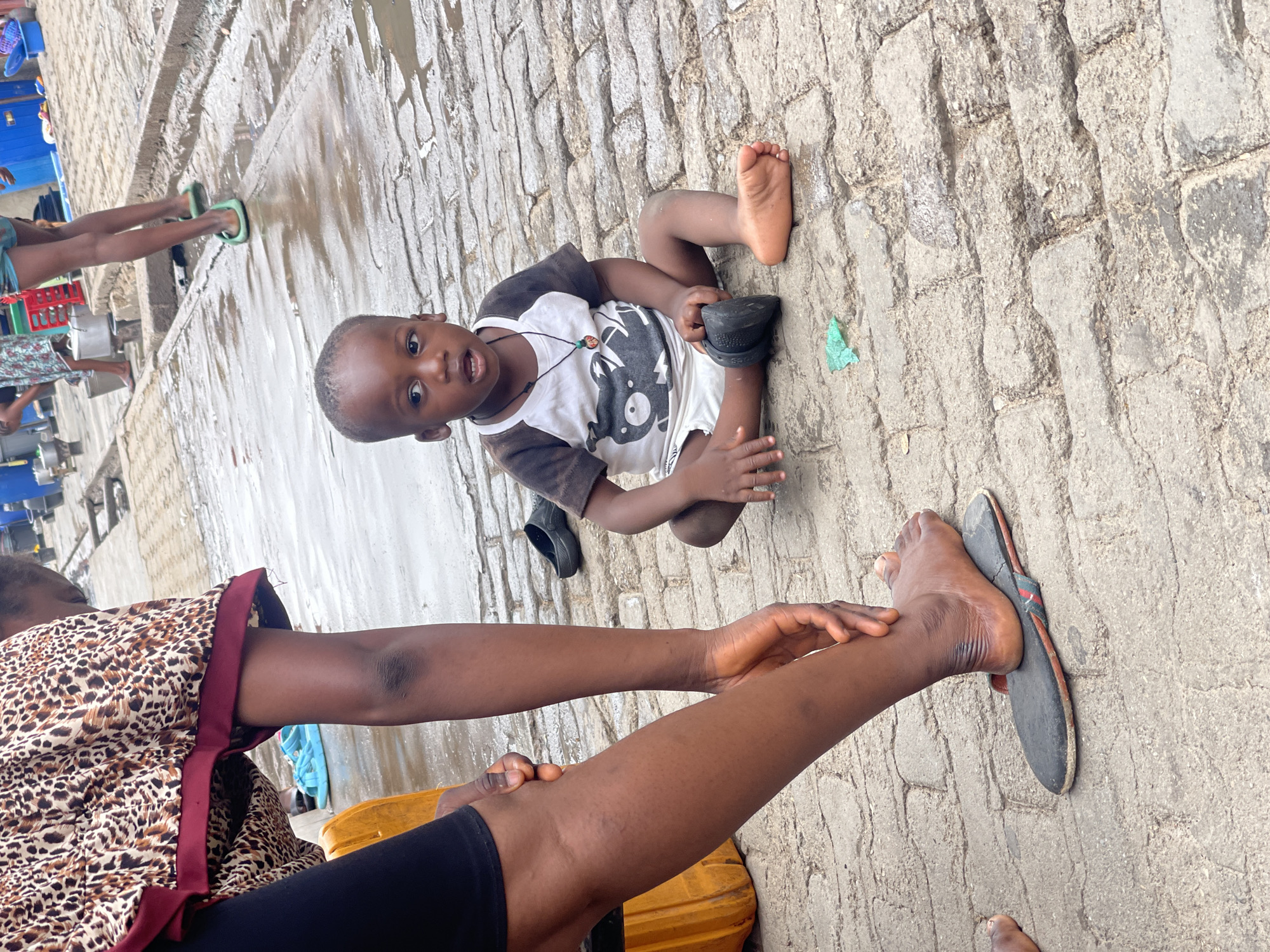 The image, set in Nyanya, Nigeria, captures a candid outdoor scene featuring a young child and parts of two adults. The ground is paved with rough, irregular grey stone blocks, marked by several reflective puddles of water, suggesting recent wetness or standing water.
In the mid-ground, a young child, likely a boy, is seated directly on the ground. He wears a white and black short-sleeved t-shirt with a cartoon-like graphic and white shorts, along with a beaded necklace. He looks directly at the camera with wide, curious eyes and a slightly open mouth. One black shoe, possibly a Croc-style sandal, is off and resting near his bare foot. A small piece of green plastic litter is visible on the ground near him.
In the bottom right foreground, an adult, dressed in a leopard-print patterned top with dark red trim and black pants, is partially visible. Her arm and hand are resting on her own leg, and her bare foot with a simple black slipper is also visible. Further in the background on the left, another person's legs wearing green flip-flops can be seen standing near what appear to be various blue and red plastic containers. The overall lighting suggests it is daytime. No readable text is visible in the frame.