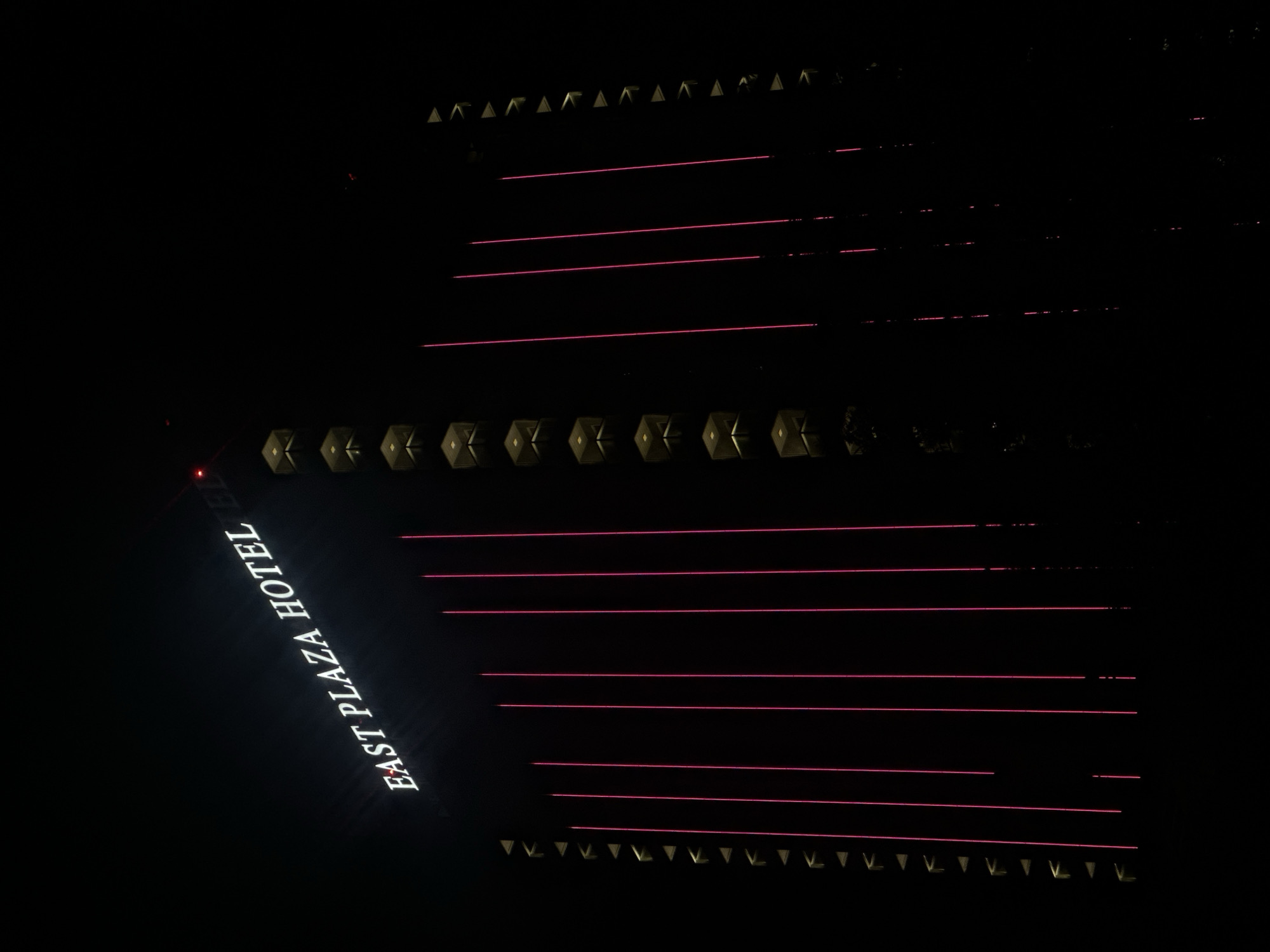 The image depicts the East Plaza Hotel at night, illuminated by its neon signage and decorative pink lighting. The hotel, located in Sofia, Bulgaria, is a tall, modern building characterized by its dark facade and distinctive vertical light elements. The "EAST PLAZA HOTEL" sign is prominently displayed in white, illuminated lettering at the top of the building. Below the sign, vertical pink lights run down the sides of the hotel, creating a striking visual effect against the dark night sky. Geometric, gold-colored architectural elements are visible along the left side of the building, interspersed with the pink lights. The overall impression is one of a vibrant, urban nightscape.