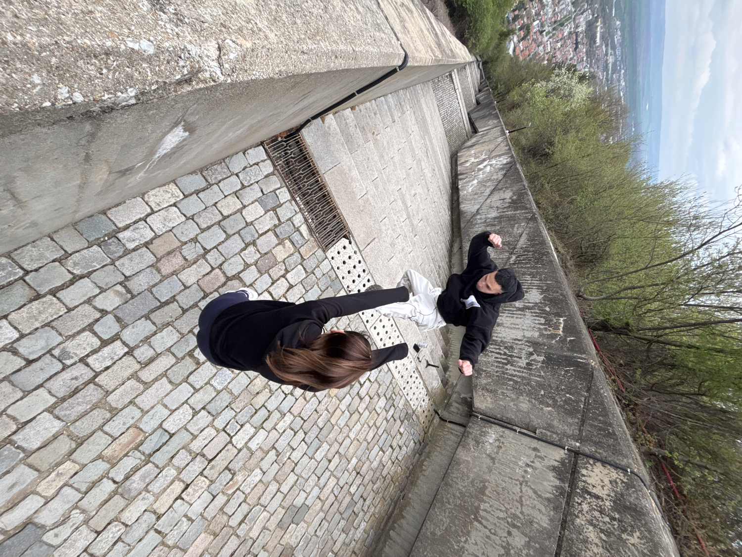 A high-angle, tilted photograph captures two individuals on a steep, narrow outdoor passage composed of cobblestones and concrete. In the foreground, a female with long brown hair, wearing a black top, is partially visible from the back, appearing to ascend the path. Further up and to the right, a male wearing a black hoodie and light-colored pants is seated or resting against a concrete wall, looking towards the camera with his fists clenched.
The passage is bordered by high concrete walls on both sides. To the right of the male figure, the concrete wall gives way to dense green trees and bushes, behind which a sprawling town or city is visible in the distant valley, under a partly cloudy or overcast sky. Metal grates are integrated into the cobblestone path near the base of the concrete wall. The overall composition strongly emphasizes the steep incline of the structure, which shows signs of weathering on its concrete surfaces. The scene appears to be set during daytime with mild weather. No visible text is present in the image.