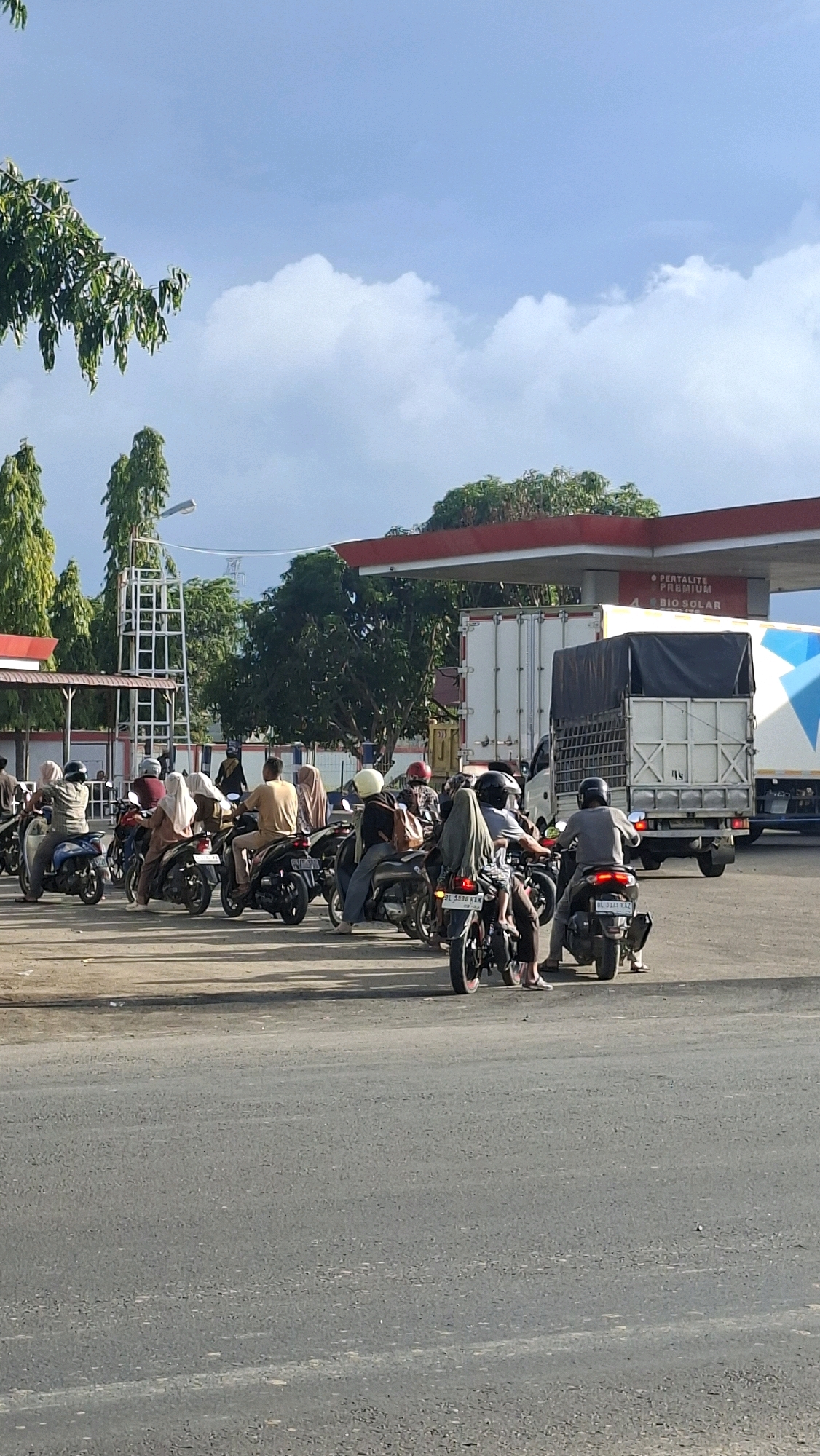 The image depicts a scene at a gas station in Keudebayu, Indonesia, with a line of motorcycles and a truck. The sky is blue with scattered clouds. A red and white gas station canopy extends over the area. Several people are riding on motorcycles, some are wearing helmets and headscarves. A large white truck with a black tarp is parked in the background. Trees are visible behind the gas station. The foreground shows an asphalt road.