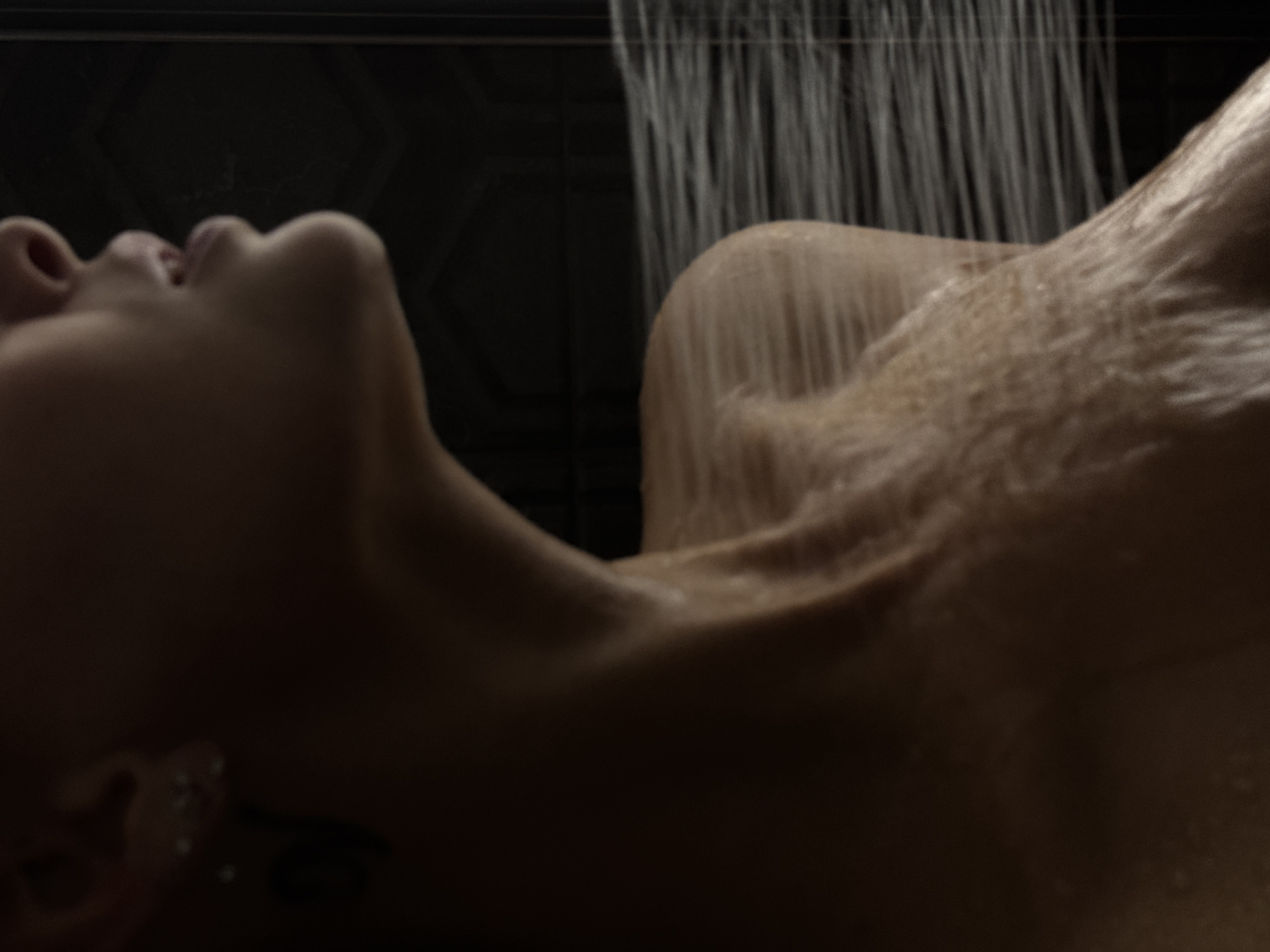The image shows a close-up of a person's upper body, from the neck up, in a shower. Water is spraying across the shoulder and upper chest, indicating the subject is being rinsed. The lighting is low, creating a dramatic effect, with shadows obscuring much of the face and body. The background is dark and indistinct, possibly a shower wall or curtain. There is no information in the image to suggest its location in Chaikovsky, Russia.
