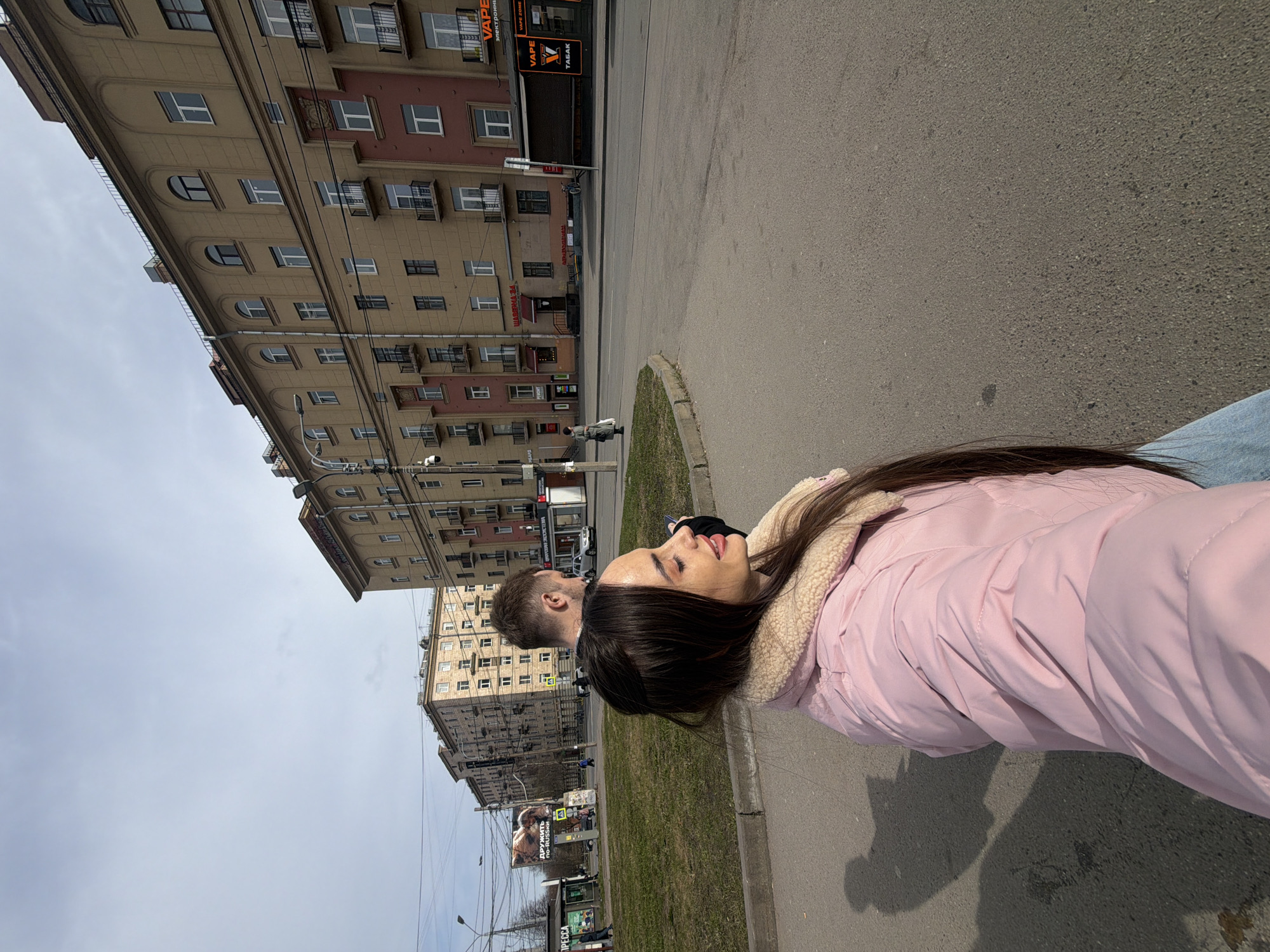 The image shows a young woman in a pink jacket walking down a street in Saint Petersburg, Russia. She is accompanied by a man. The background includes multi-story buildings, power lines, and signage in Russian.