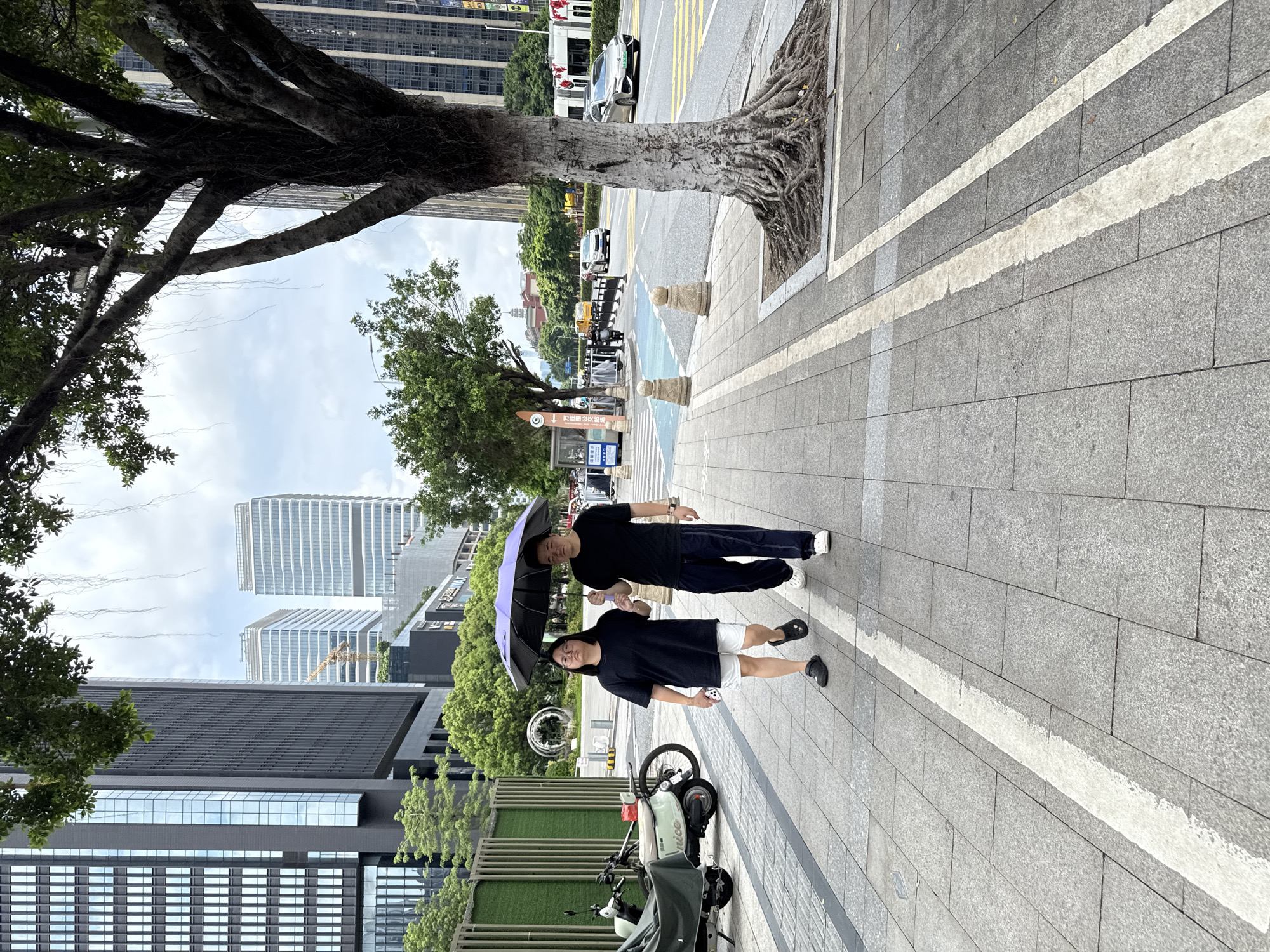 A man and a woman are walking on a sidewalk under a purple umbrella in Haizhu District, China. The woman is wearing a black t-shirt, white shorts, and black sandals. She is holding her phone in her left hand. The man is wearing a black t-shirt and dark pants with white shoes. They are walking side-by-side, with the man holding the umbrella. The sidewalk is paved with gray tiles and bordered by a white line. To the left of the sidewalk, there are some parked scooters and a green fence. In the background, there are modern buildings, trees, and a street with cars. The sky is cloudy.