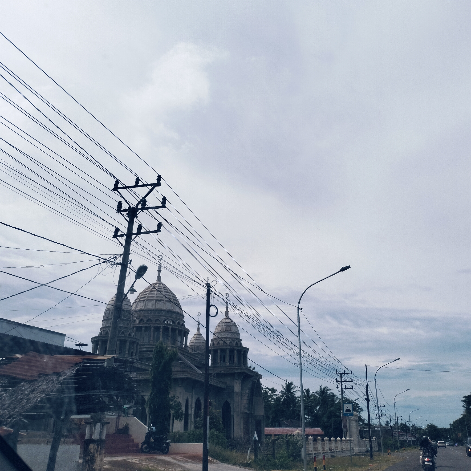 This image captures a vibrant street scene on an overcast day in Teungoh Baro, Indonesia, featuring a distinctive mosque, extensive power lines, and daily street activity.
In the midground on the left, a large, grey building with multiple domed structures stands prominently. These domes, adorned with intricate patterns and topped with crescent moon finials, clearly identify it as a mosque. The building's material appears to be concrete or stone, giving it a weathered or possibly unfinished look. A tall, slender tree grows in front of the mosque, and a motorcycle is parked nearby.
The upper and central parts of the frame are heavily crisscrossed with numerous black power lines, which converge at a substantial utility pole located in the mid-left. Additional utility poles and curved streetlights extend along the right side of the road, receding into the distance.
On the right side, a paved road is visible, with several motorcycles and a car in the background, indicating active traffic. Individuals are seen riding the motorcycles. The roadside is lined with abundant tropical vegetation, including tall palm trees, characteristic of the region.
The sky is a uniform, light grey, heavily overcast with visible cloud formations, suggesting a cloudy day. The overall lighting is soft and diffused. The photograph appears to be taken from the perspective of someone inside a vehicle, looking out onto the street. No discernible text is visible on any signs or structures.
