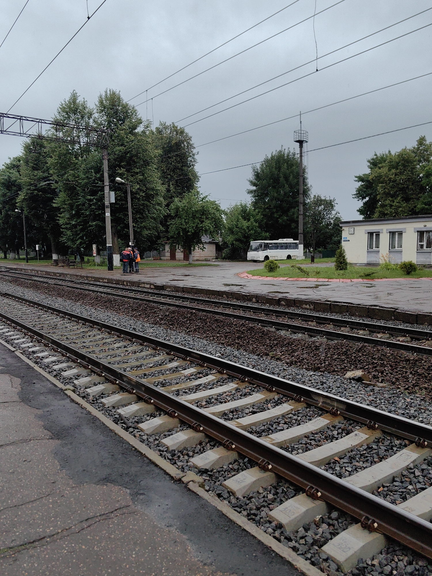 The image depicts a view of a train station in Fanipal, Belarus. In the foreground, two sets of train tracks run parallel to each other, composed of gravel ballast and concrete ties. The tracks are wet, suggesting recent rain. To the left of the tracks is a paved platform, also wet. In the middle ground, a group of three individuals wearing high-visibility vests are gathered near a utility pole. One of the individuals is partially obscured by another. Beyond them, a row of trees lines the area, with a white bus parked on a paved surface to the right. Further to the right is a light-colored building with windows, and a tall, slender tower with a cage-like structure at its top. Overhead, multiple power lines are visible against a cloudy sky. The overall scene is overcast and appears to be a functional, albeit somewhat bleak, railway environment.