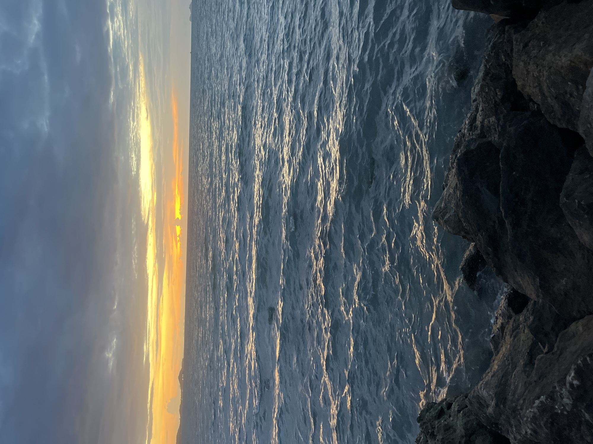 A wide shot captures a body of water during a sunset in Baan Plai Laem, Thailand. The sky is a gradient of dark gray clouds at the top transitioning to a bright yellow and orange band near the horizon. The sun is partially obscured by clouds, casting a warm glow. The water is a deep blue-green with visible ripples and highlights of golden light reflecting the sunset. In the foreground, dark, textured rocks line the bottom of the frame, creating a rough shoreline. The horizon shows a distant landmass, possibly islands or a coastline, with faint outlines of structures. The overall scene depicts a serene and natural coastal environment at dusk.
