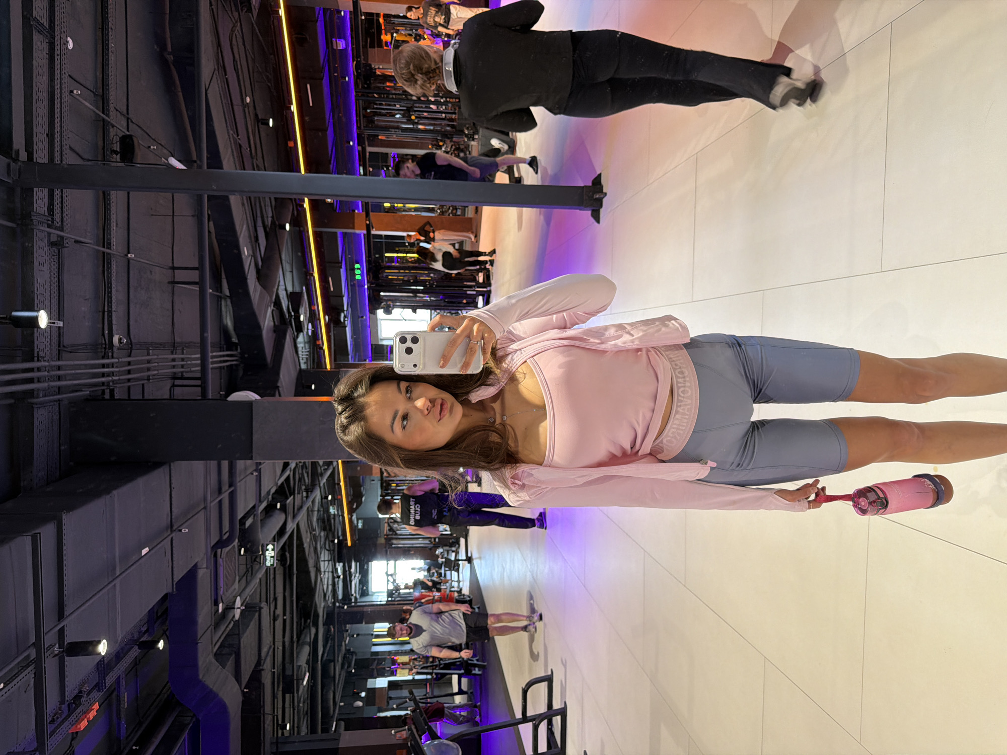A woman takes a selfie in a gym setting with people in the background. She is wearing a pink top and gray athletic shorts, holding a pink water bottle. The gym has weights, weight machines, and purple lighting. The location is not specified in the image, but may be the city of Krasnoyarsk, Russia.
