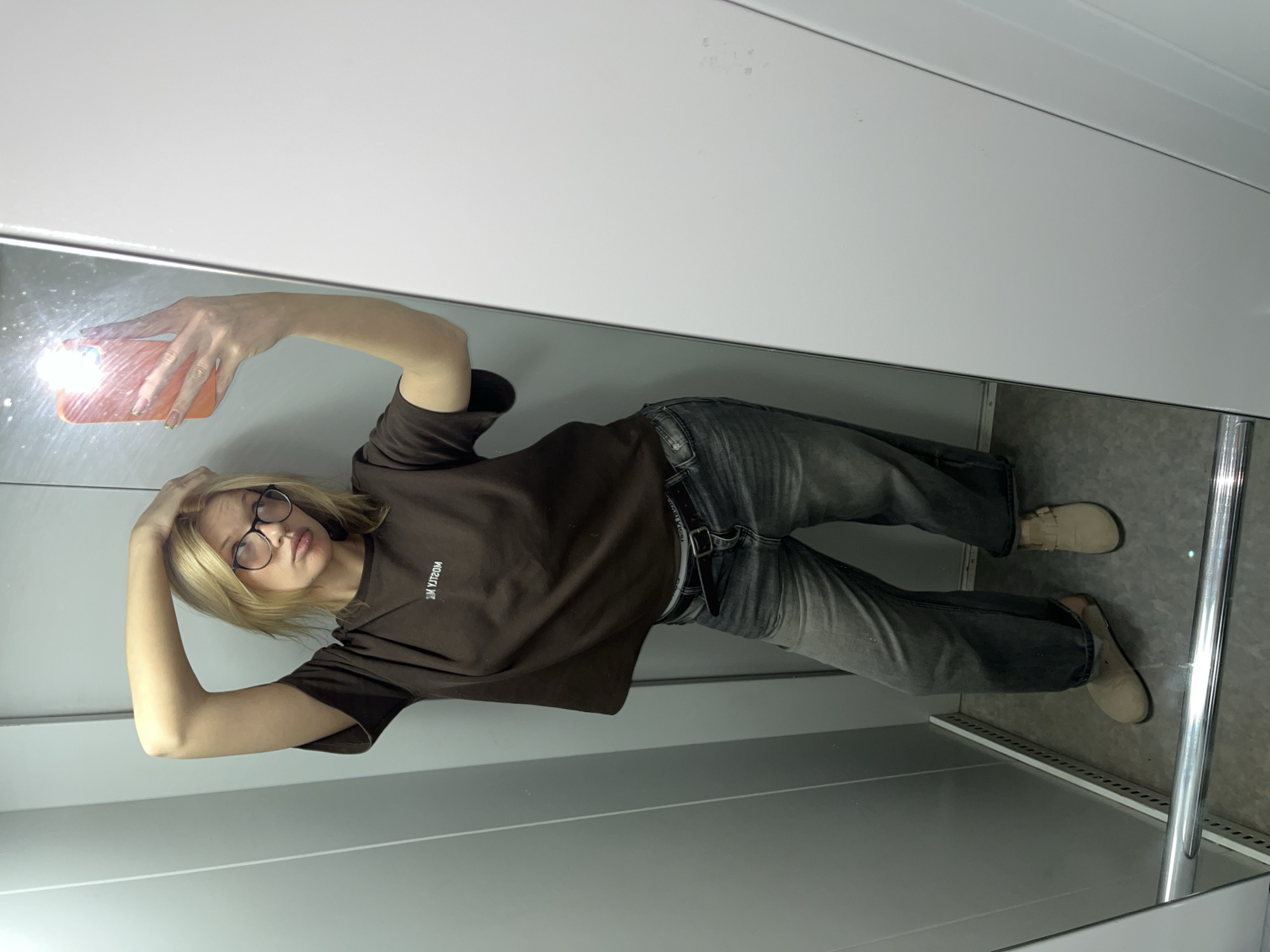 A woman with blond hair and glasses is taking a selfie in a mirror. She is wearing a brown t-shirt, jeans, and tan shoes. She is holding a cell phone in her left hand. The environment appears to be inside a building with white or light gray walls. The location may be Chaikovsky, Russia.