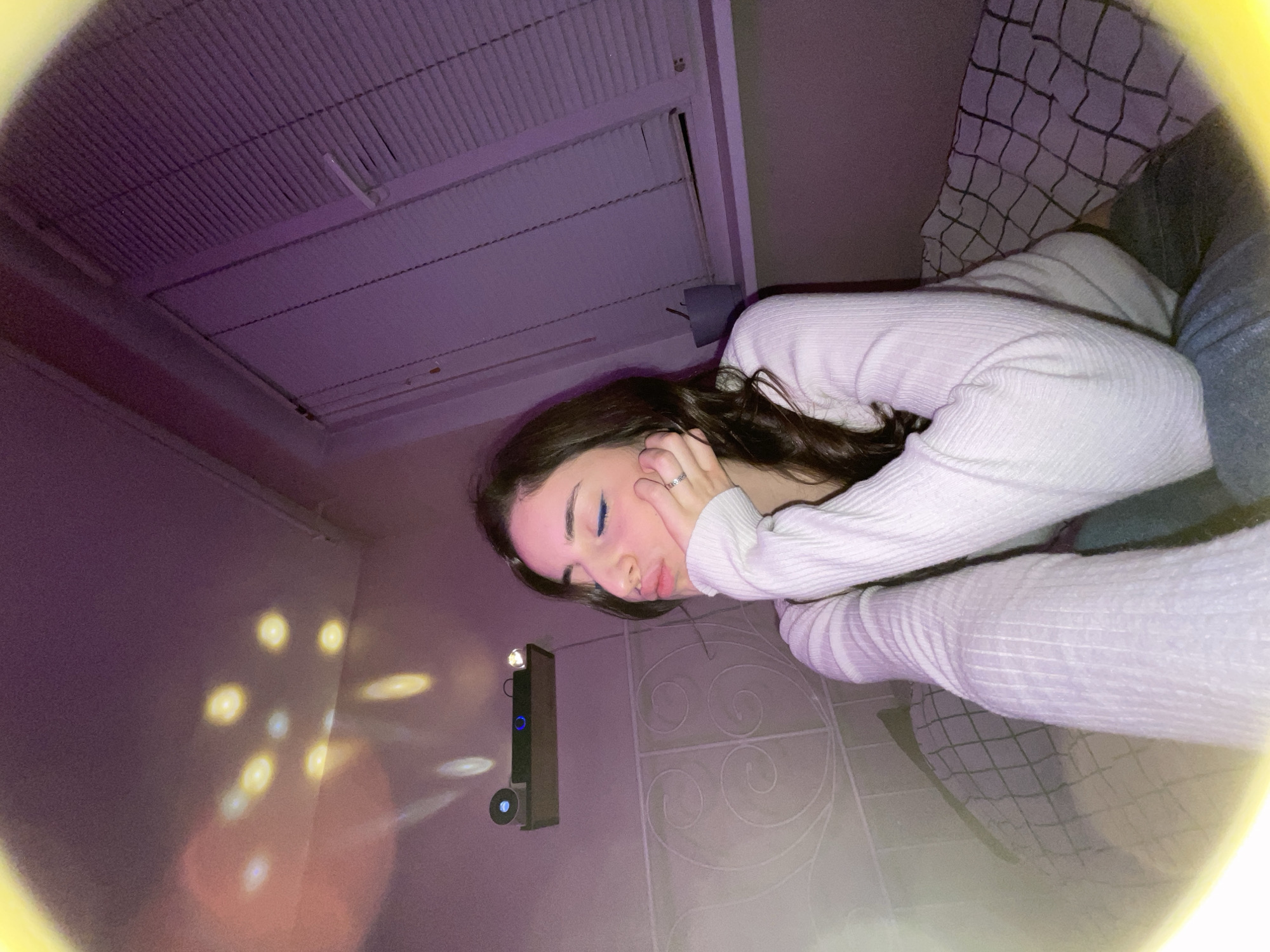 A young woman is reclining on a bed in a room with a low ceiling. She is wearing a white, long-sleeved shirt and appears to be resting her head on her hand. The room is illuminated by purple lighting, and there are light patterns projected on the wall from a projector that is mounted on the wall. The background includes a ceiling vent and a bed with a patterned blanket. The location in the city of Moscow, Russia is not discernible from the given image.