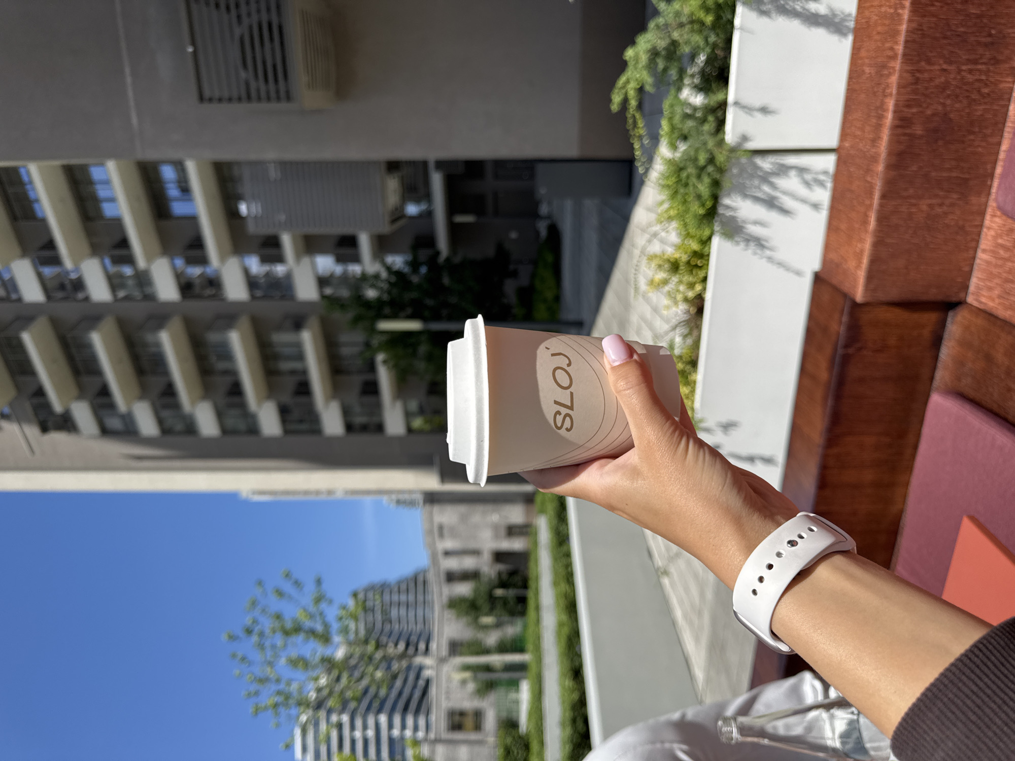 A person holds a "SLOJ" brand coffee cup in an urban setting, as seen through the background of a multi-story building complex and trees in Belgrade, Serbia. The person wears a white smartwatch and has pink nail polish. The coffee cup is positioned in the foreground, and the buildings and greenery provide a background. An outdoor seating area with wooden-style furniture is on the right-hand side of the frame.