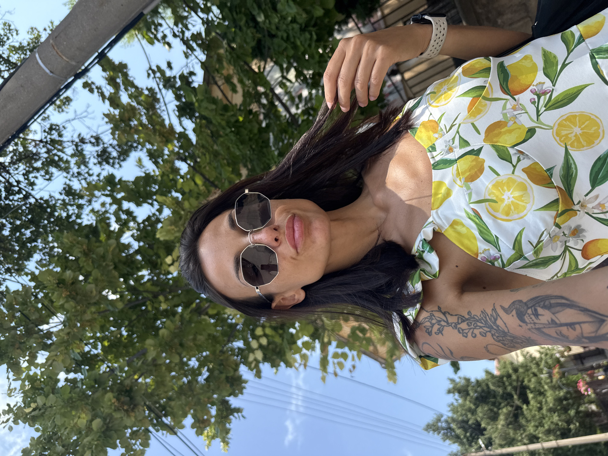 The media file is a close-up, vertically oriented selfie of a young woman with long, dark hair and sunglasses. She is wearing a white top with a vibrant lemon print. Her right arm is visible, featuring a tattoo that appears to be a stylized portrait with floral elements. She is touching her hair with her left hand, and her expression is relaxed. The background consists of green foliage and a clear blue sky with some clouds, suggesting an outdoor setting on a bright, sunny day. The lighting indicates it is likely daytime. Visible elements in the background include a large pipe or beam above and to the left of the subject, and what appear to be power lines in the lower portion of the frame. There is no visible text. The location context provided is Pleven, Bulgaria.