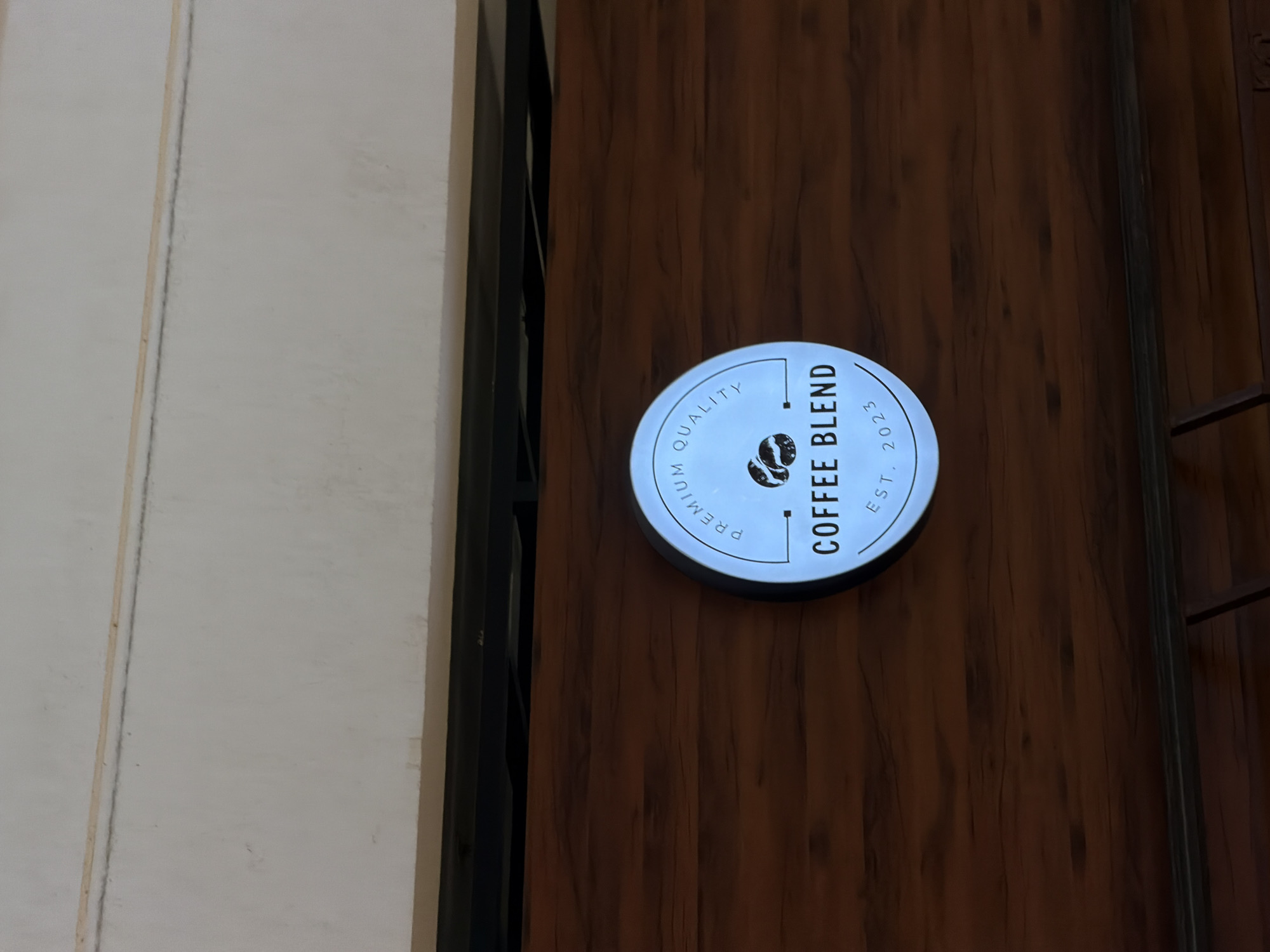 The image shows a close-up of a circular, illuminated sign with text and a graphic. The sign is mounted on a wooden wall, which has a visible grain pattern. The sign itself is white with black lettering and graphics. At the top of the circle, it reads "PREMIUM QUALITY" in an arc. In the center of the sign are two stylized coffee beans. Below the coffee beans, in larger font, it reads "COFFEE BLEND". At the bottom of the circle, it reads "EST. 2023" in an arc. The background above the wooden wall appears to be a white ceiling or upper wall with a darker horizontal line running across it. This image does not provide any information about the city of Sharm El Sheikh, Egypt.