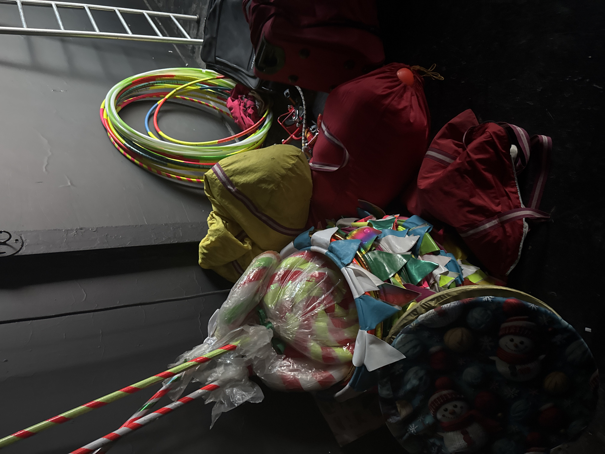 In Aksu, Turkey, the image depicts a dimly lit room, likely a storage area or backstage, containing various props and equipment. Prominently featured are several colorful hula hoops stacked against a dark gray wall. To the left, a cluster of items are wrapped in clear plastic, including what appear to be large, striped lollipops or candy canes, and a smaller, cylindrical object. A decorative banner with triangular flags in shades of blue, white, green, and pink is draped over some of the items. Also visible are several large, fabric bags, one yellow and two red, some appearing to be duffel bags or sacks. A black suitcase or bag is situated to the right of the hula hoops, and another red bag with straps is in the foreground. A round, fabric item with a festive snowman and snowflake pattern is on the far left, suggesting a possible holiday or event theme. The overall impression is one of disarray, with items piled together, hinting at preparation for or aftermath of an event. A silver ladder is partially visible in the background, leaning against the wall.