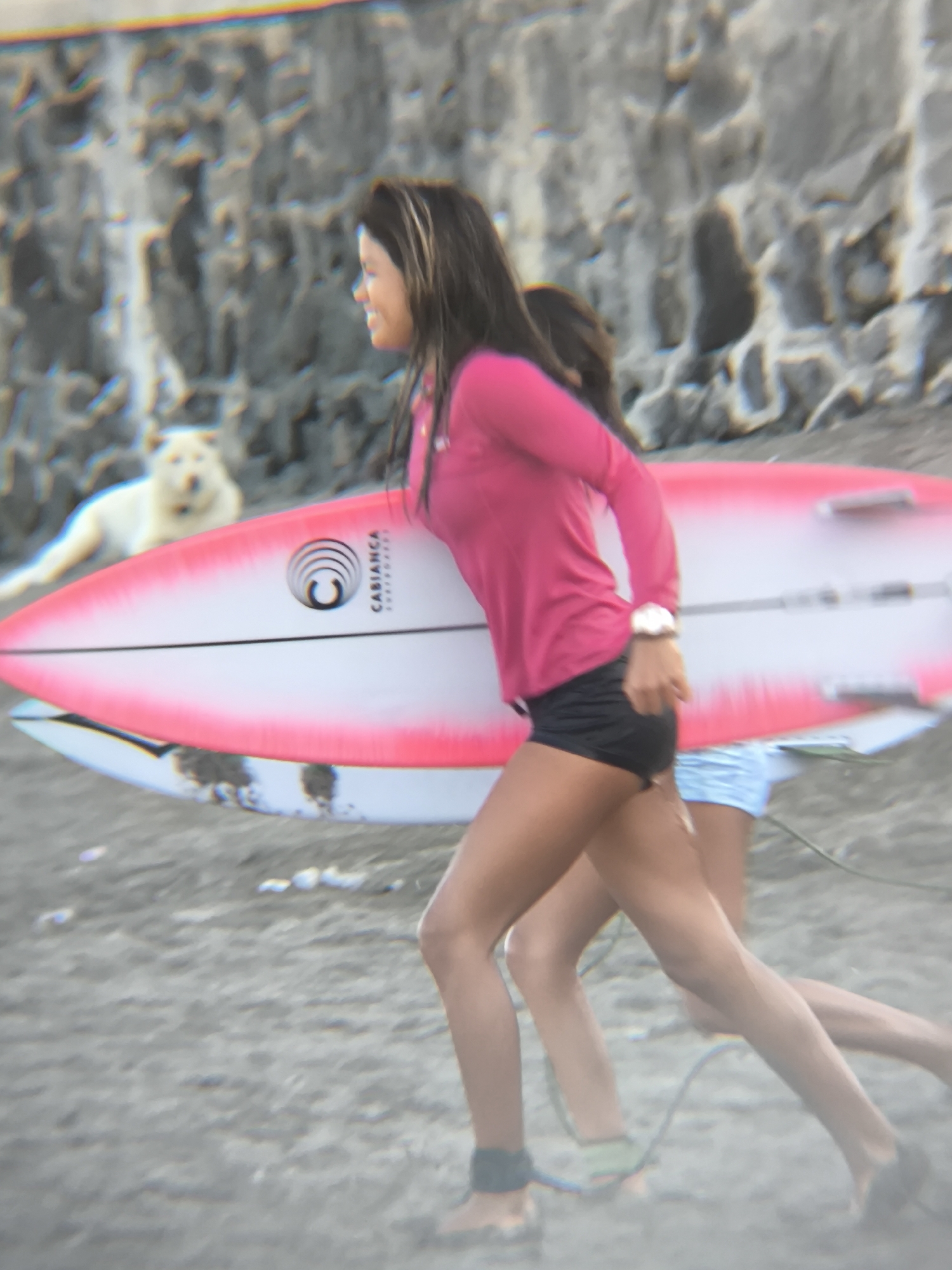 The photo shows two women running on a beach carrying surfboards. The first woman is wearing a pink long-sleeved shirt and black shorts. The other woman is partially obscured. Both are barefoot and have leashes attached to their ankles. Behind them, a white dog is lying down. The background includes a rocky cliff face. The surfboards are white with pink edges and a logo that includes the word "CABIANCA". The location is North Kuta, Indonesia, based on the style of surfboards and the environmental features.