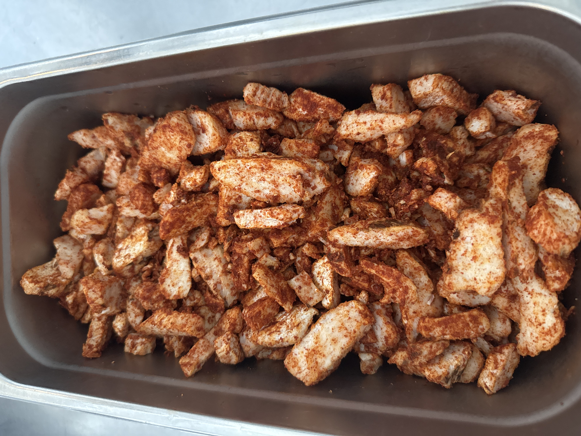 A top-down shot captures a rectangular stainless steel container filled with seasoned, chopped pieces of white meat, likely chicken. The meat is coated in a reddish-brown spice blend, giving it a slightly textured appearance. The container is situated on a surface that appears to be a stainless steel countertop, with a blurred, light-colored background suggesting an indoor setting, possibly a kitchen or food preparation area. The lighting is neutral, casting minimal shadows.