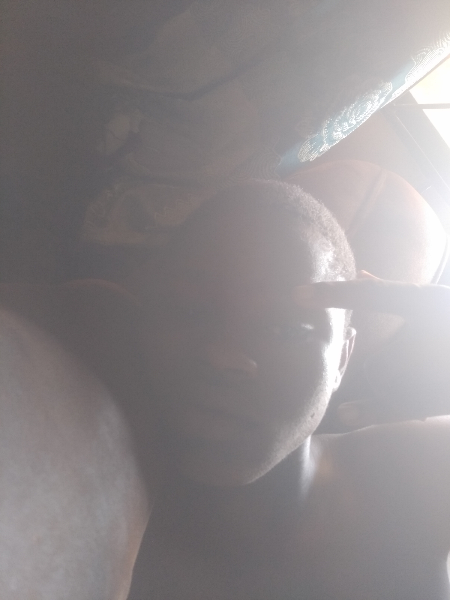 A person is taking a selfie indoors with strong, harsh lighting. The person has short, dark hair and appears to be touching their forehead with their hand. The location is not possible to determine from this image.