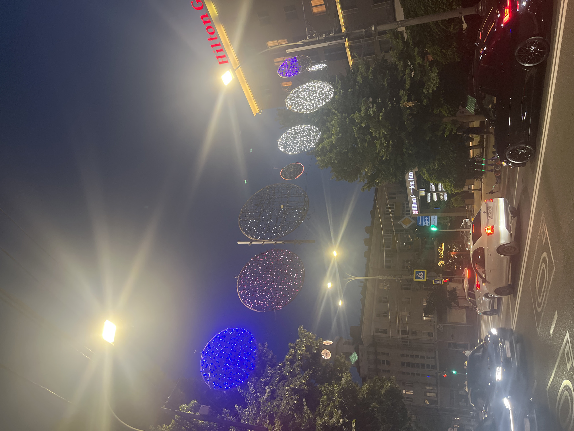 The image depicts an urban street scene at night, likely in Krasnodar, Russia, given the provided context. The main subjects are several cars, including a black car in the foreground and a white car further down the street. There are also decorative light installations hanging above the street, which appear to be large, circular or oval shapes adorned with numerous small lights. Some of these are illuminated with white lights, while one prominently features blue lights, and another has purple lights.
The setting is an urban environment with multi-story buildings visible on either side of the street. Trees with dense foliage are interspersed along the sidewalk and near the buildings. The sky is dark, indicating it is nighttime. A street lamp with a bright, diffused light is visible on the left side of the frame, contributing to the overall illumination of the scene.
The activity taking place is not explicitly evident, but the presence of cars on a street suggests normal traffic. The decorative lights suggest a festive occasion or an effort to enhance the ambiance of the street, possibly for holidays or a special event.
Notable details include the "Hilton Garden Inn" sign clearly visible on one of the buildings, which helps establish a specific location context. Another sign with Cyrillic text, reading "24-23:00" which likely indicates opening hours, is also present. Pedestrian crossing signs are visible, indicating a crosswalk. The time of day is clearly night, and the weather appears clear. There are no clear indications of emotions from any visible people, as they are not a primary focus.
