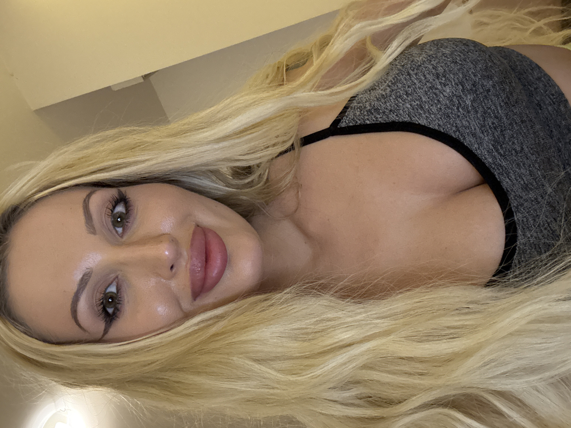 A close-up, head-and-shoulders portrait shows a fair-skinned blonde woman with long, wavy hair. She is wearing a grey sports bra with black trim. Her eyes are green and she has dark eyeliner and long eyelashes. Her lips are full and painted with a pinkish-brown lipstick. She is looking directly at the camera. The background is a plain, light-colored wall. The image is taken indoors.