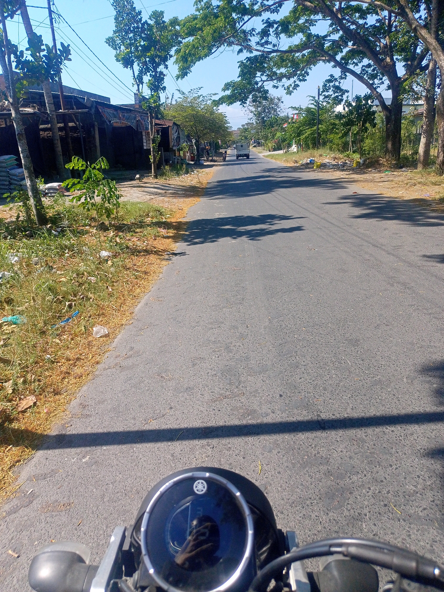 The image depicts a street scene in Renteng, Indonesia, from the perspective of someone riding a motorcycle. The asphalt road stretches into the distance, flanked by vegetation and buildings. On the left side of the road, there are stacks of bags, likely cement, and a rudimentary structure with a corrugated metal roof. Trees with dense green foliage line both sides of the road, casting shadows on the pavement. A small white vehicle is visible in the distance, and a motorcycle is parked on the right side of the road. The sky is clear and blue, indicating a sunny day. The foreground shows the handlebars and instrument cluster of the motorcycle, including a circular digital display with a Yamaha logo. Scattered trash is visible along the roadside.