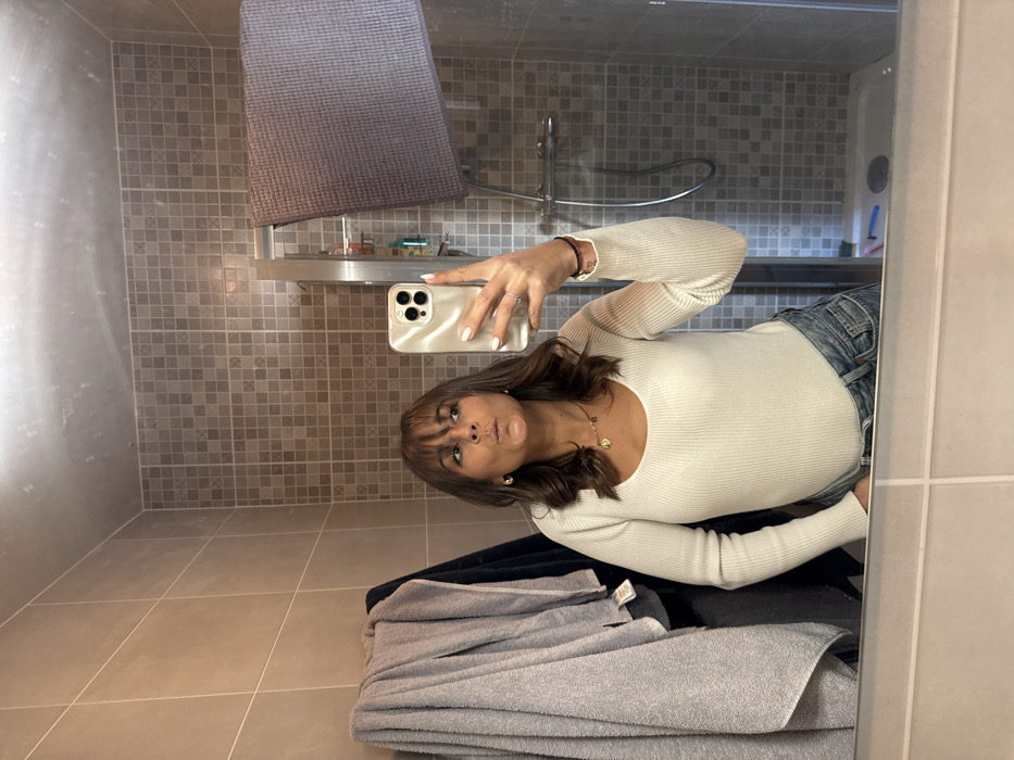The scene depicts a woman taking a mirror selfie in a bathroom. The specific location in Cannes, France, cannot be confirmed from the visual evidence within the image.
**People:** One female individual is present, reflected in the mirror. She has medium-length brown hair with bangs and is making a puckered lip expression, looking towards her smartphone.
**Subjects/Objects (on the person):** She is wearing a light-colored, possibly cream or off-white, ribbed long-sleeved top with a scoop neck, tucked into light blue denim jeans. She has a gold-colored necklace with two pendants, gold-colored hoop earrings, and a ring on the ring finger of her right hand. Her fingernails are painted white. She is holding a smartphone with a light-colored (champagne or gold) case in her right hand, aimed at her reflection. A dark band or bracelet is visible on her left wrist.
**Environment:** The bathroom features a large rectangular mirror, reflecting the entire scene. The walls are tiled; an upper section displays small, square, light brown or beige mosaic-style tiles, while a lower section shows larger rectangular beige tiles. To the left of the woman's reflection, several grey and one dark blue or black towel are hanging on a fixture. To the right, a shower enclosure with a glass door and metal frame is visible. Inside the shower, a showerhead, mixer tap, and several bottles are discernible on a shelf. A dark grey or purple towel is draped over a metal rail attached to the shower's glass door. The floor of the shower appears to be a light-colored tray. A portion of a sink or counter is visible at the bottom edge of the mirror.
**Actions/Interactions:** The woman is actively engaged in taking a photograph of herself using her smartphone's front-facing camera, captured via reflection in the mirror. Her pose and facial expression are directed towards this action.