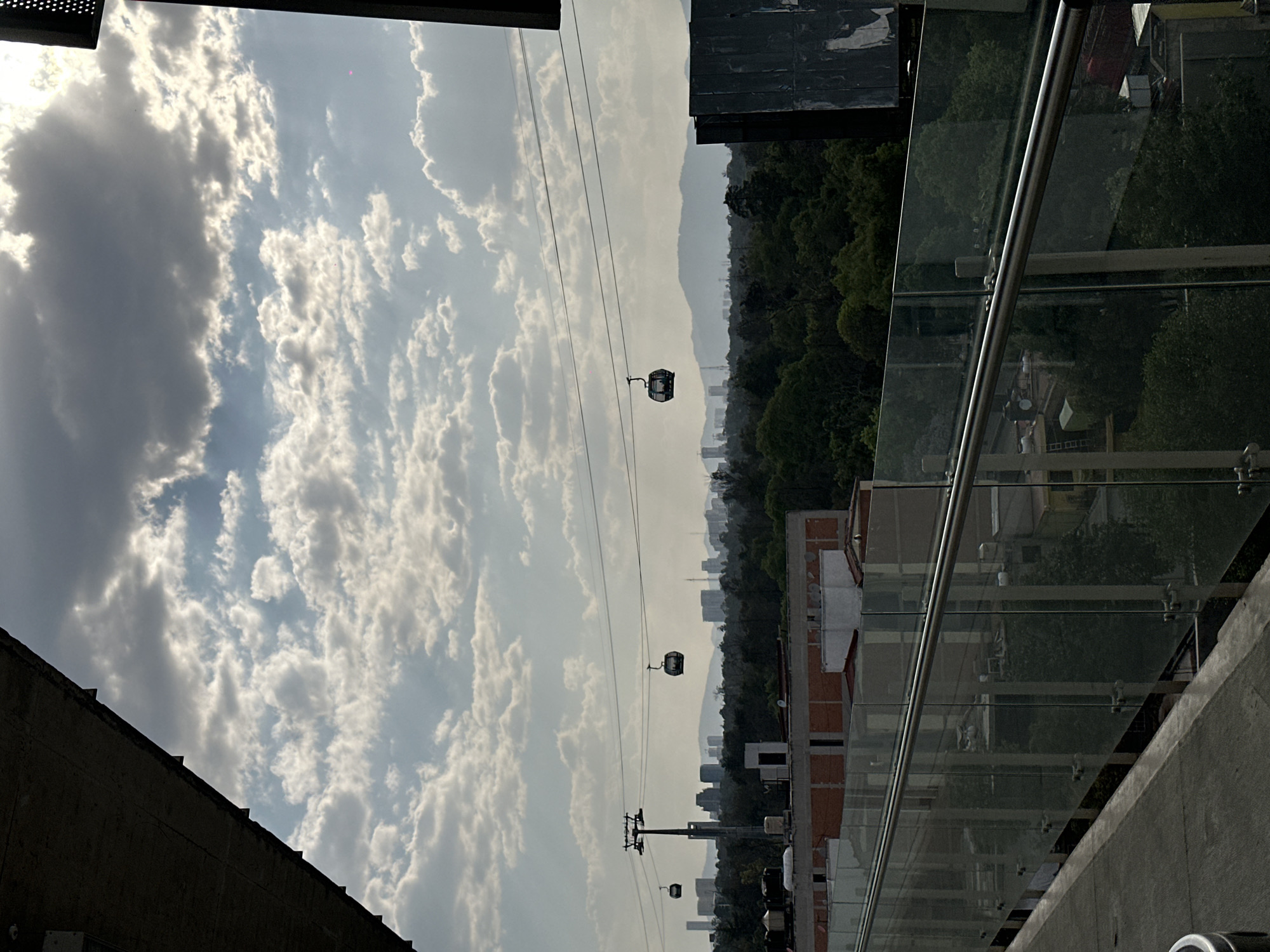 This media shows an elevated outdoor view of an urban landscape, likely from a building or viewing platform. The scene is dominated by a cloudy sky, with bright white cumulus clouds contrasted by darker gray cloud masses. The sun, though not directly visible, appears to be illuminating the clouds from the upper left.
Suspended across the mid-ground are multiple cable cars, or gondolas, on overhead wires. At least two cable car cabins are clearly visible, dark in color, moving or stationary against the sky. One cable car pylon, supporting the wires, is also present towards the lower middle.
Below the cable car lines, a dense green forest or park area stretches across a ridge, transitioning into a hazy urban sprawl with numerous distant buildings. Further in the background, light-colored, layered mountains or hills define the horizon, obscured by atmospheric haze.
In the immediate foreground, to the right, there is a modern architectural structure featuring a glass barrier with a metal handrail, suggesting a balcony or terrace. A concrete wall is visible beneath the railing, and parts of buildings with red-tiled or reddish-brick roofs are seen further down. On the far left, a dark, angled architectural element, possibly part of a building's roof or facade, frames the scene.
The lighting suggests it is daytime, likely late morning or afternoon, under partly cloudy skies. The extensive cable car system over a mountainous, hazy city points to a location like Mexico City, Mexico, which features such infrastructure. No people or visible text are discernible in the image.