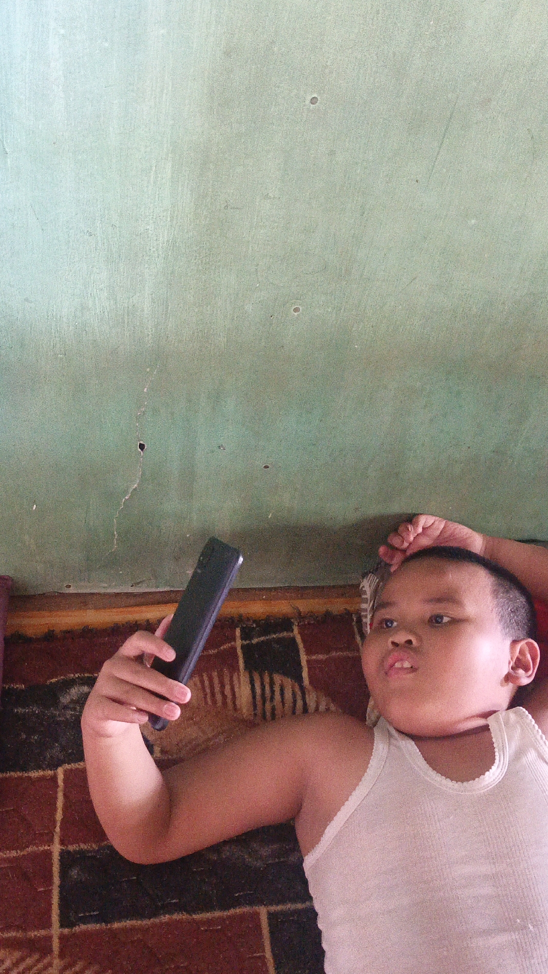 In Sungai Rasau, Indonesia, a young boy in a white tank top lies on his back on a patterned rug, looking at a dark smartphone held in his left hand. He is positioned near a wall painted a light green, which shows signs of wear, including cracks and small holes. The boy's right arm is bent at the elbow, with his hand resting near his head. The lighting suggests an indoor environment.