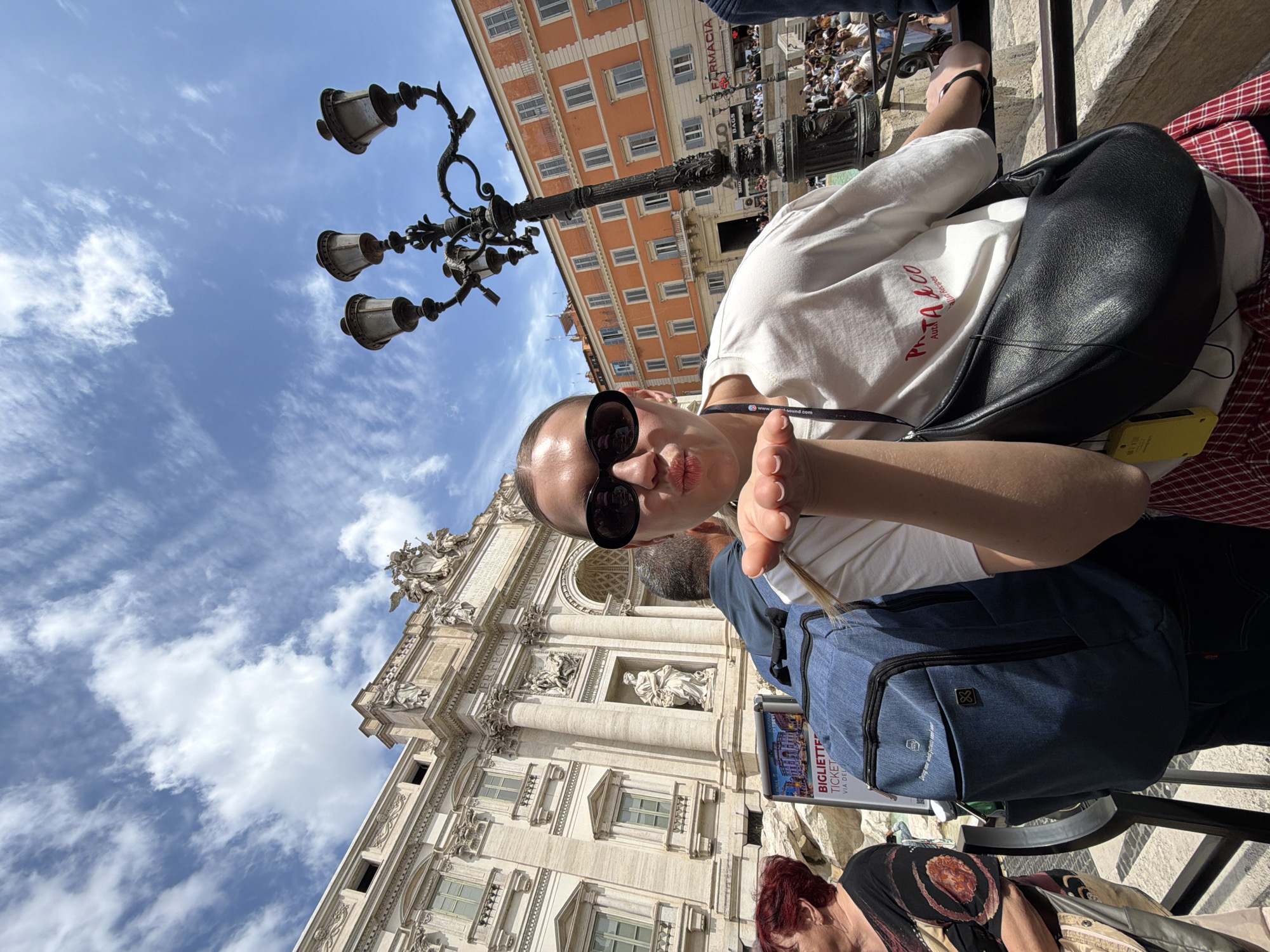 This image captures a lively selfie taken in front of the iconic Trevi Fountain in Rome, Italy, during daytime with a clear blue sky and scattered white clouds.
The primary subject, a woman, is positioned in the foreground, wearing round black sunglasses and a white t-shirt with red text, partially visible as "Pieta & Co.". She has a black shoulder bag and a lanyard displaying "www.next-sound.com". She is looking directly at the camera with a playful, puckered-lip expression, her hand slightly extended. Behind her, another person's head and shoulder are partially visible, and further to the bottom right, a person with reddish-brown hair can be seen.
The grand Baroque facade of the Trevi Fountain dominates the background, showcasing its intricate sculptures, classical columns, and statues. Across from the fountain, in the mid-ground, stands an orange-hued building with numerous windows and a visible "FARMACIA" sign. A crowd of people is gathered around the fountain and in the square, indicating a popular tourist destination. An ornate black street lamp is prominently featured in the upper left portion of the image. Additionally, a blue backpack with the text "bring your world everywhere with you" and a logo is visible, along with a "BIGLIETTERIE TICKET" sign. A small yellow device, possibly a battery pack or audio guide, is attached to the main subject's black bag. The overall atmosphere suggests a sunny and busy tourist experience.