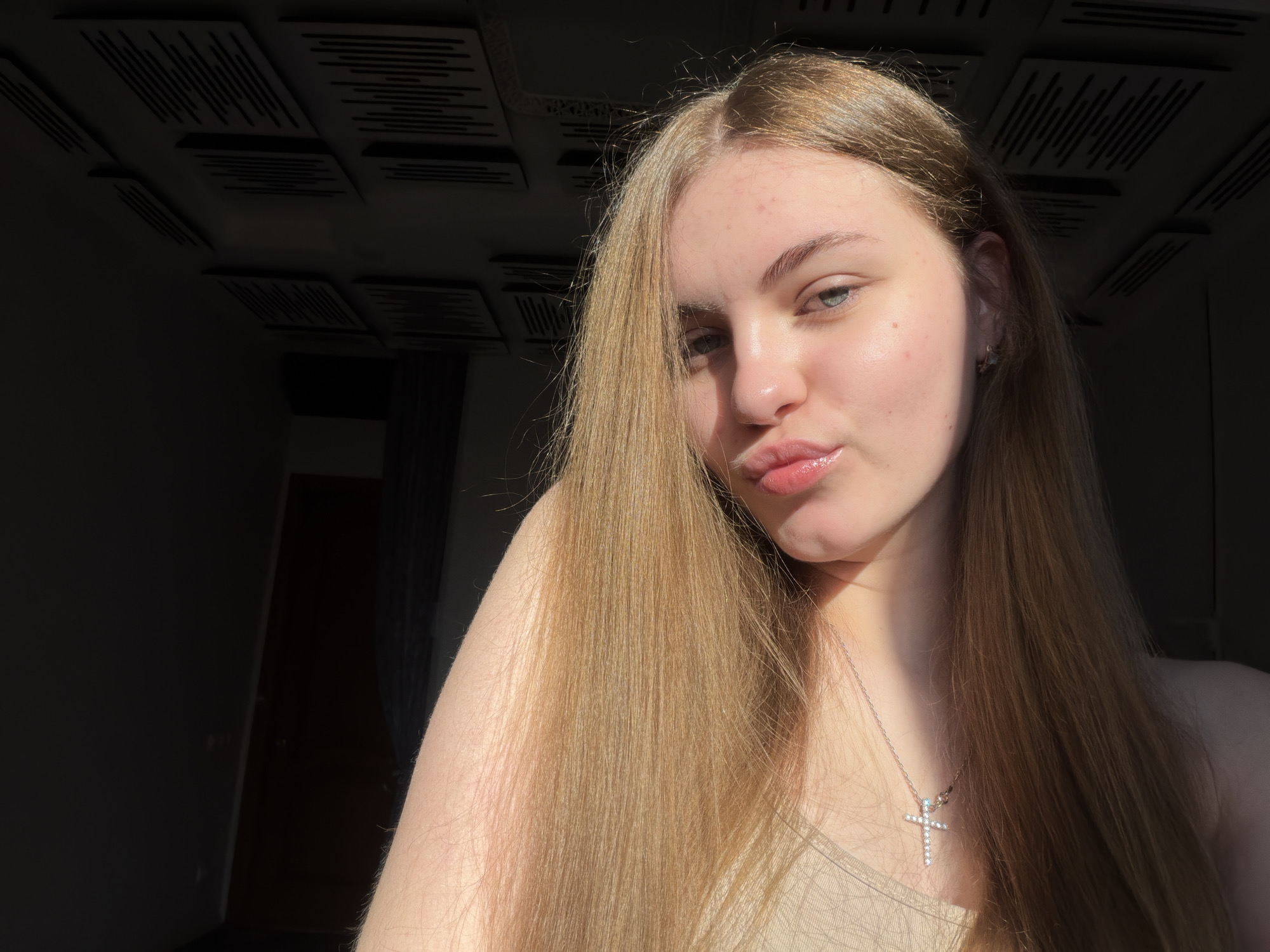 A young woman with long, blonde hair and light blue eyes is captured in a close-up portrait. She is wearing a tan-colored top and a silver necklace with a cross pendant. Her lips are slightly parted as if she is about to speak or kiss. The lighting suggests the sun is shining on her face from the right, casting a warm glow. The background appears to be an indoor setting, possibly a room with dark walls and a ceiling adorned with square acoustic panels. The overall mood of the image is intimate and serene, with the woman's gaze directed towards the camera. This image was taken on a sunny afternoon in an apartment located in the city of Dnipro, Ukraine.