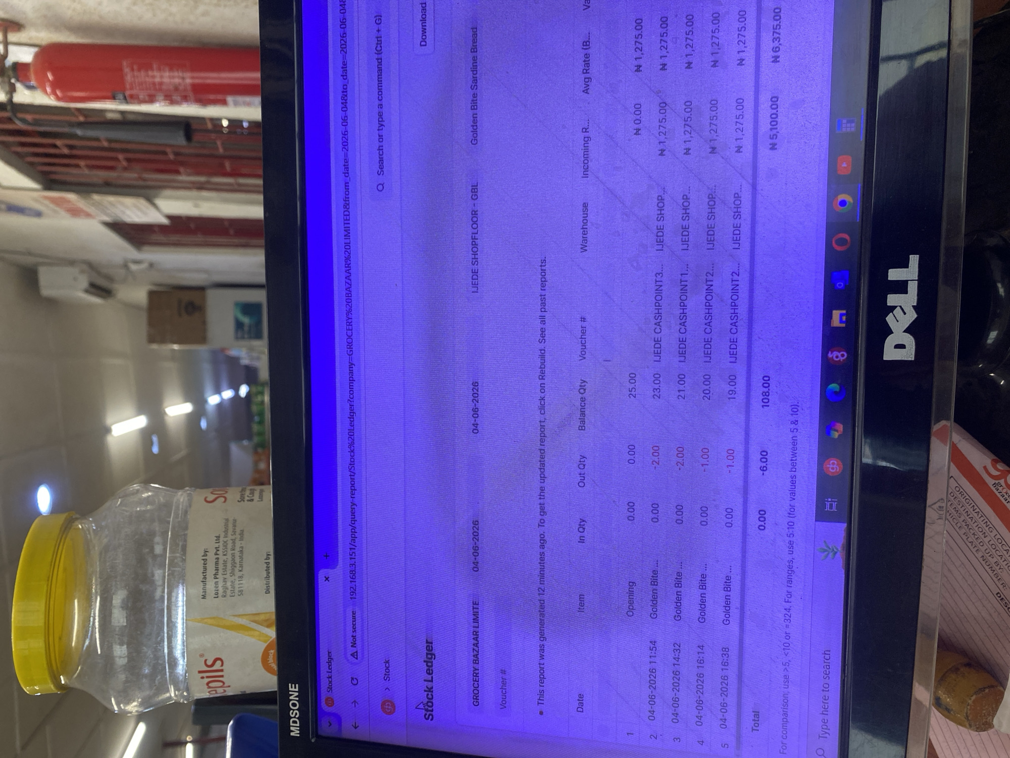 An angled shot captures a computer monitor displaying a "Stock Ledger" report, with various objects in the foreground and background, in an indoor setting in Ikorodu, Nigeria. No people are visible.
The monitor, a DELL brand, shows a web browser interface with a "Stock Ledger" report for "GROCERY BAZAAR LIMITED," dated "04-06-2026." The report details stock movements for "Golden Bite Sardine Bread" at "IJEDE SHOPFLOOR - GBL" and "IJEDE CASHPOINT" locations. Columns include "Date," "Item," "In Qty," "Out Qty," "Balance Qty," "Voucher #," "Warehouse," and "Val." The report indicates an opening balance of 25.00 units, followed by several "Out Qty" transactions totaling -6.00, resulting in a final balance of 19.00 for the listed items. Monetary values are displayed in Nigerian Naira (N), with amounts like N 1,275.00 and totals of N 5,100.00 and N 6,375.00. A message states the report was "generated 12 minutes ago."
In the foreground, a clear plastic jar with a yellow lid is visible. Its label reads "epils," "Manufactured by: Lozen Pharma Pvt. Ltd. Raghav Estate, KSSIDC Industrial Estate, Shiggaon Road, Savanur 581118, Karnataka - India," and "Sore th & Cough Lozenge." It also mentions "Distributed by: MDSONE." In the background, a red fire extinguisher is mounted on a wall, and parts of a shelving unit or rack are discernible. A printed document in the bottom right corner shows text including "ORIGINATING LOCATIO," "DESTINATION LOCAT," and "VEHICLE PLATE NUMBER," suggesting shipping or logistics information. The overall scene indicates an office or stockroom environment focused on inventory management and record-keeping.