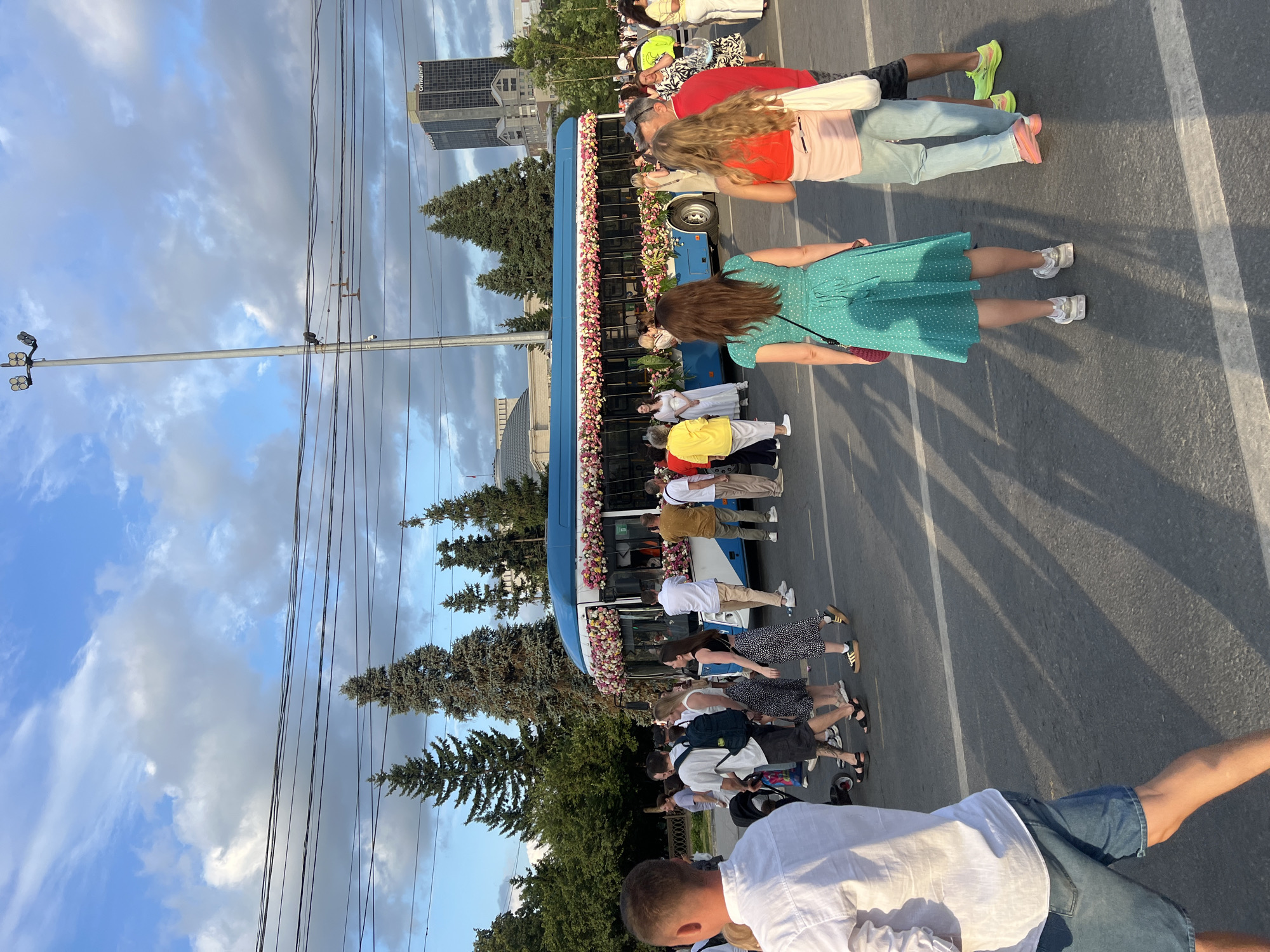 A group of people are gathered on a street next to a blue bus decorated with flowers. The bus appears to be stationary. The people are standing in various positions, some facing the bus and others facing away. The scene is outdoors, with trees and a cloudy sky visible in the background. There are also power lines and a tall building with text on it. The time of day appears to be late afternoon or early evening, given the lighting and shadows. The overall atmosphere seems relaxed, with some individuals appearing to be enjoying the event. The people are dressed in casual summer clothing. The text on the building is difficult to decipher due to the distance and angle. There is no clear indication of the specific event taking place, but it could be a parade, a festival, or a gathering of some sort.