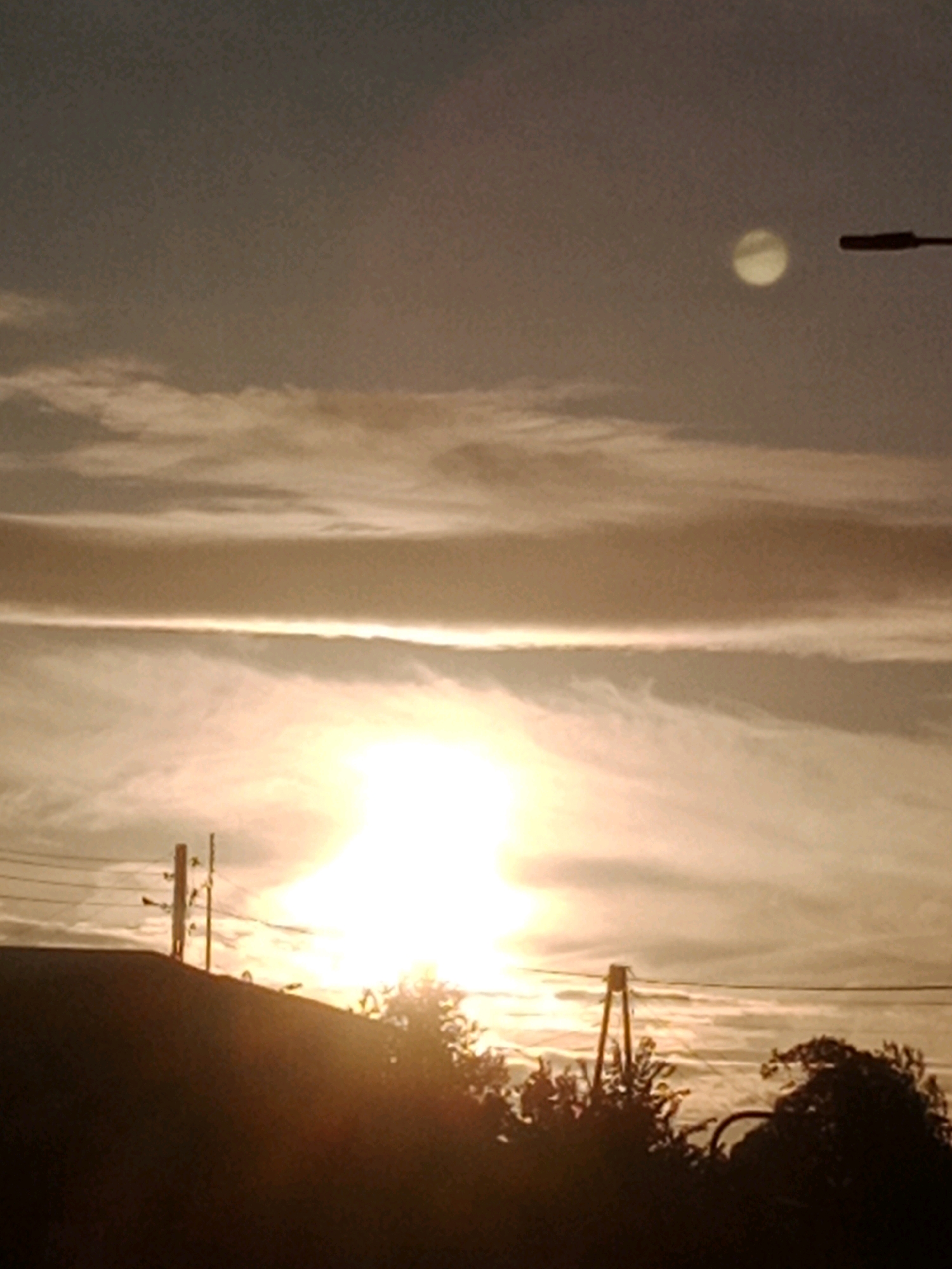 A low-angle, slightly hazy photograph captures a sunset scene. The sun, a bright, blown-out white disk, dominates the lower-middle portion of the frame, casting a warm glow. Thin, wispy clouds stretch horizontally across the sky, reflecting the sun's light. In the upper right, a small, circular, pale object, possibly the moon, is visible. A dark, horizontal silhouette of a streetlamp extends from the right edge of the frame towards the left, partially obscuring the celestial bodies. In the foreground, the dark silhouette of a rooftop occupies the bottom edge, with electrical poles and wires stretching across the scene. Trees and vegetation, also in silhouette, are visible at the bottom right. The overall impression is a dusky, urban twilight. This scene could be any urban area experiencing sunset, and the provided crops offer no specific identifiers to locate it in Bydgoszcz, Poland.
