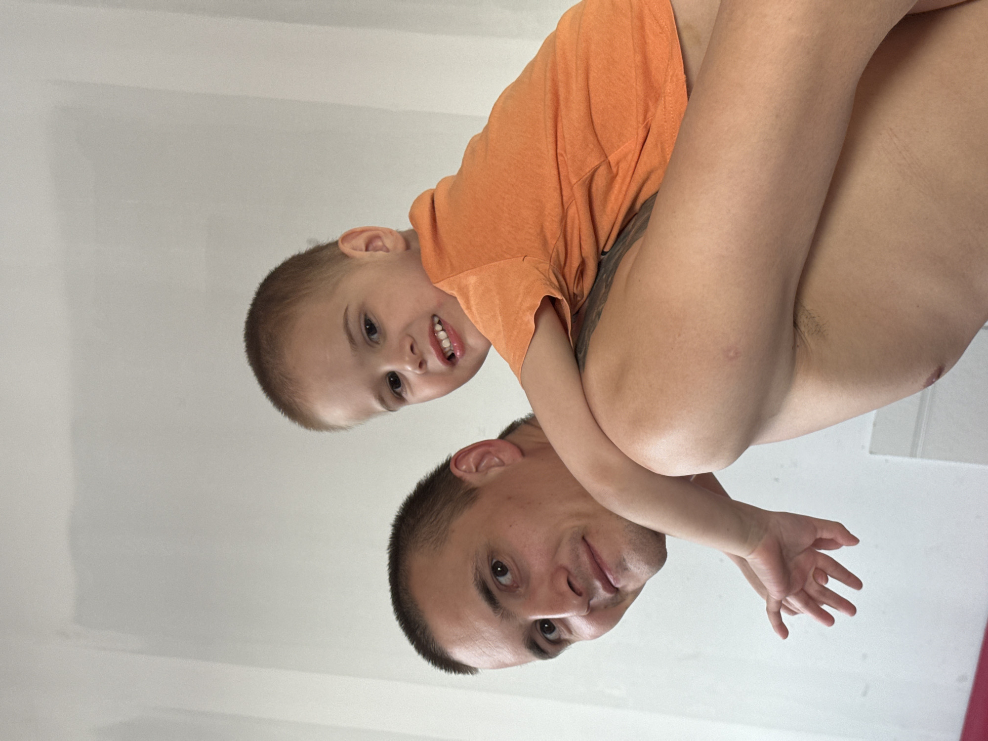 A man and a young boy are looking down at the camera. The boy is wearing an orange t-shirt and is being held by the man. The man is not wearing a shirt. They are both smiling. The background is a plain white wall. There is no indication in the image that the location is in the city of Chaikovsky, Russia.