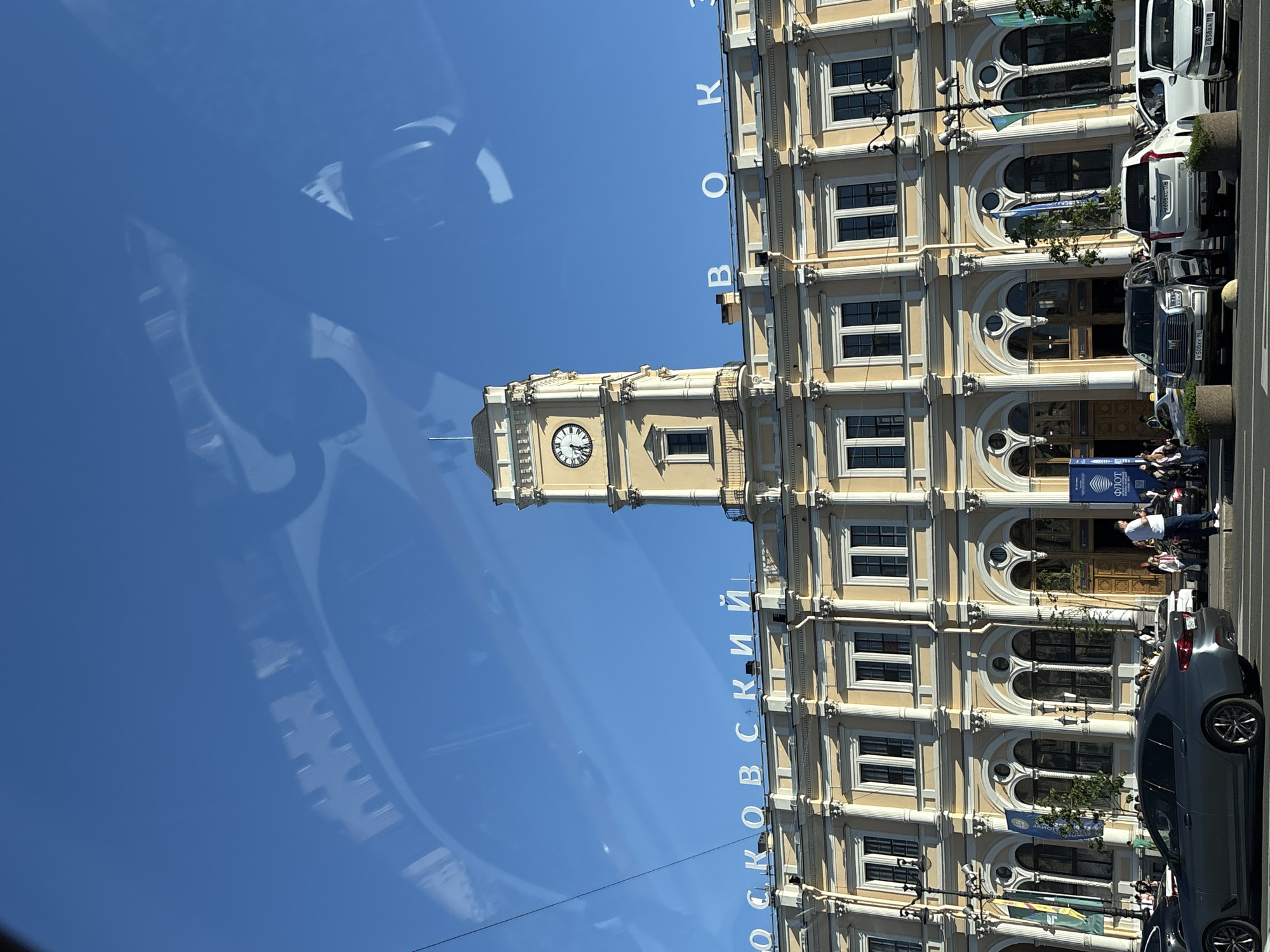 This image depicts the facade of the Moskovsky Vokzal (Moscow Railway Station) in Saint Petersburg, Russia, under a clear blue sky. The multi-story building is rendered in a classical architectural style, featuring a light yellow or cream color, numerous rectangular windows with dark frames, and a series of arched openings on the ground floor. White classical columns and decorative cornices adorn the facade. A prominent clock tower, also yellow/cream, is positioned on the left side of the building, displaying a clock face indicating approximately 12:45. Below the clock, a small balcony is visible. White capital letters spelling "ОСКОВСКИЙ ВОКЗ" are mounted on the roofline, identifying the "Moskovsky Vokzal."
At street level, a variety of vehicles are present, including a dark gray sedan (BMW), a silver SUV (Mitsubishi Outlander), a dark gray SUV (Haval H9), a white SUV (Cadillac Escalade), and a white sedan (Volkswagen Polo), among others. Most vehicles appear to be parked or stopped along the curb. Numerous pedestrians are visible on the sidewalk and congregating near the building's entrances, engaged in typical urban activities such as walking and standing. One man in a white shirt and dark pants stands on the street next to a white SUV. Cylindrical concrete planters or barriers are placed along the street. Several vertical banners, including blue and green ones, hang between the arched ground-floor openings. Reflections of a car's interior, possibly from the vehicle the photograph was taken from, are noticeable in the upper portion of the clear blue sky. The scene is brightly illuminated by sunlight.