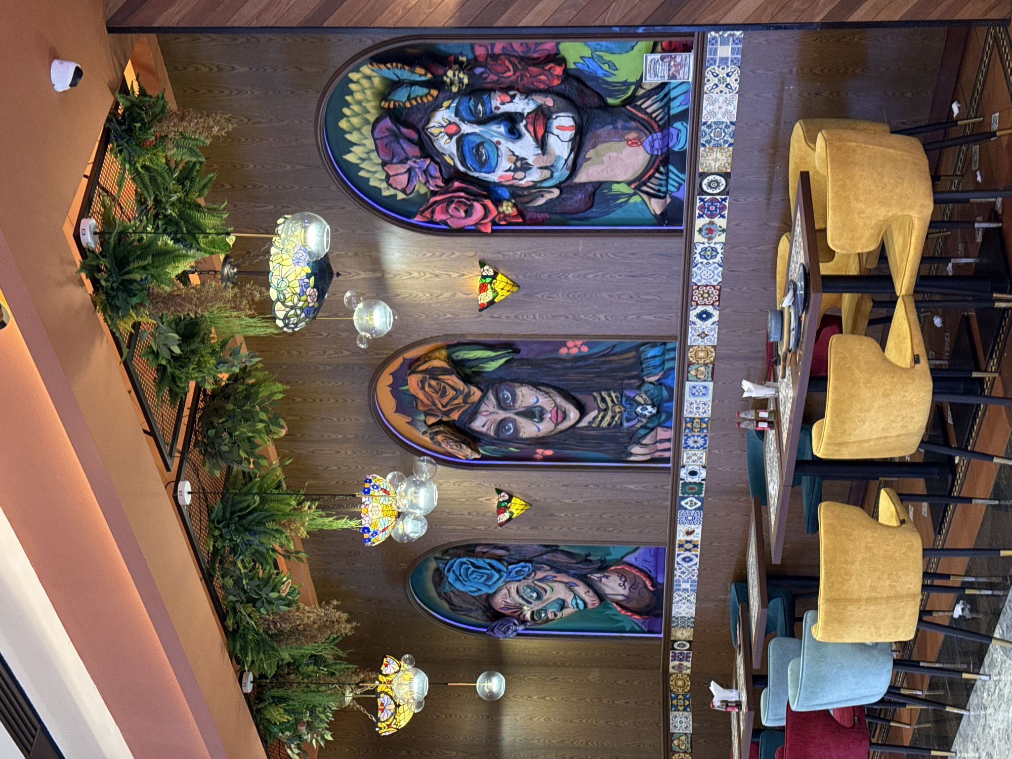 The image shows the interior of a restaurant in Kuala Lumpur, Malaysia. The focus is on the ceiling adorned with three murals depicting La Catrina, a symbol associated with the Day of the Dead. The murals are set within arches, each illuminated with neon blue lighting, and accompanied by decorative butterfly-shaped lights.
Adjacent to the murals is a tiled strip with a pattern of blue, brown, and beige, running vertically down the frame. Below this is a row of tables with a variety of chairs in red, blue, green, and mustard yellow.
The upper-left corner of the image shows a section of the wall decorated with greenery, and lighting fixtures with colorful stained glass shades.