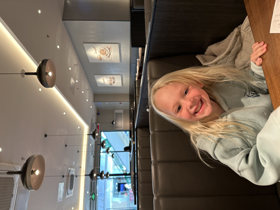 A young girl with long blonde hair sits at a table in a restaurant. She is wearing a light blue sweatshirt and smiling at the camera. The restaurant has a modern design with a long row of booths and pendant lights hanging from the ceiling. Pictures are displayed on the wall. A glimpse of a shopping mall entrance with escalators is visible through the restaurant's window.