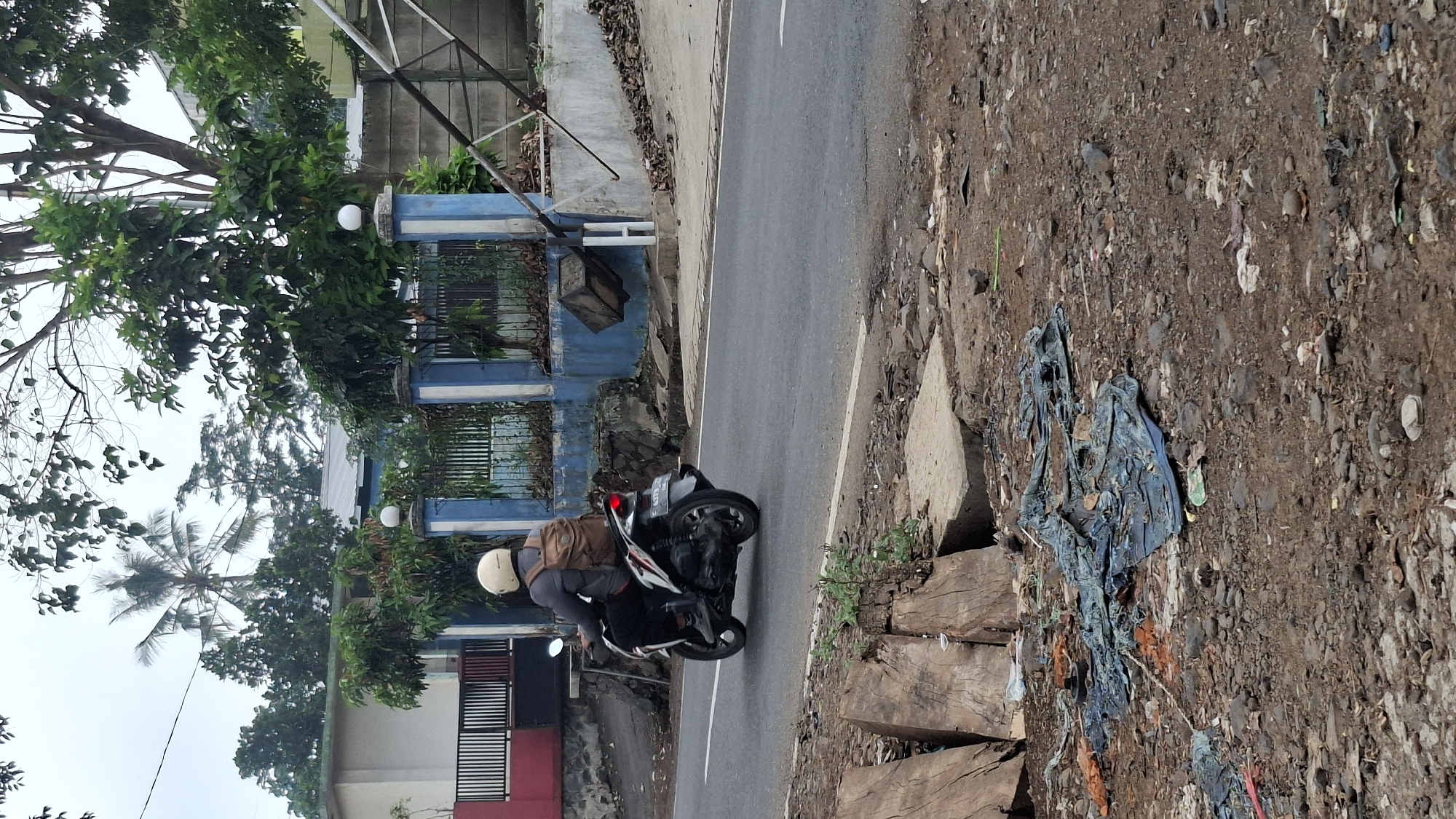 A person wearing a light-colored helmet and a tan backpack is riding a dark-colored scooter on a paved road. The scooter has a license plate with the number B 4331 ZAX. The rider is wearing a gray long-sleeved shirt and dark pants. In the background, there is a blue and white fenced area with pillars and a house with a red section and black railing. Trees, including a palm tree, are visible. The foreground shows a rocky and uneven ground with scattered debris, including pieces of wood and what appears to be blue tarp. The overall scene depicts a roadside environment in a potentially semi-urban or rural setting, possibly in Cilaku, Indonesia, given the context.