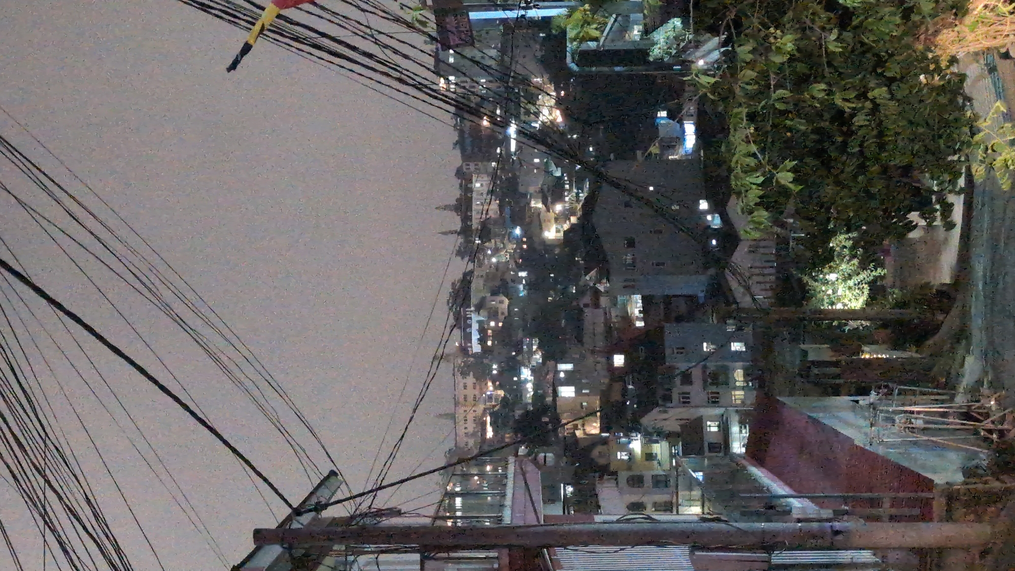 The image shows an urban scene at night in Da Lat, Vietnam. The camera is positioned looking out over a hillside city. The night sky is overcast. Buildings with lights on dot the landscape. The foreground shows power lines and nearby roofs, including corrugated iron. The area in the extreme lower right is covered in foliage.