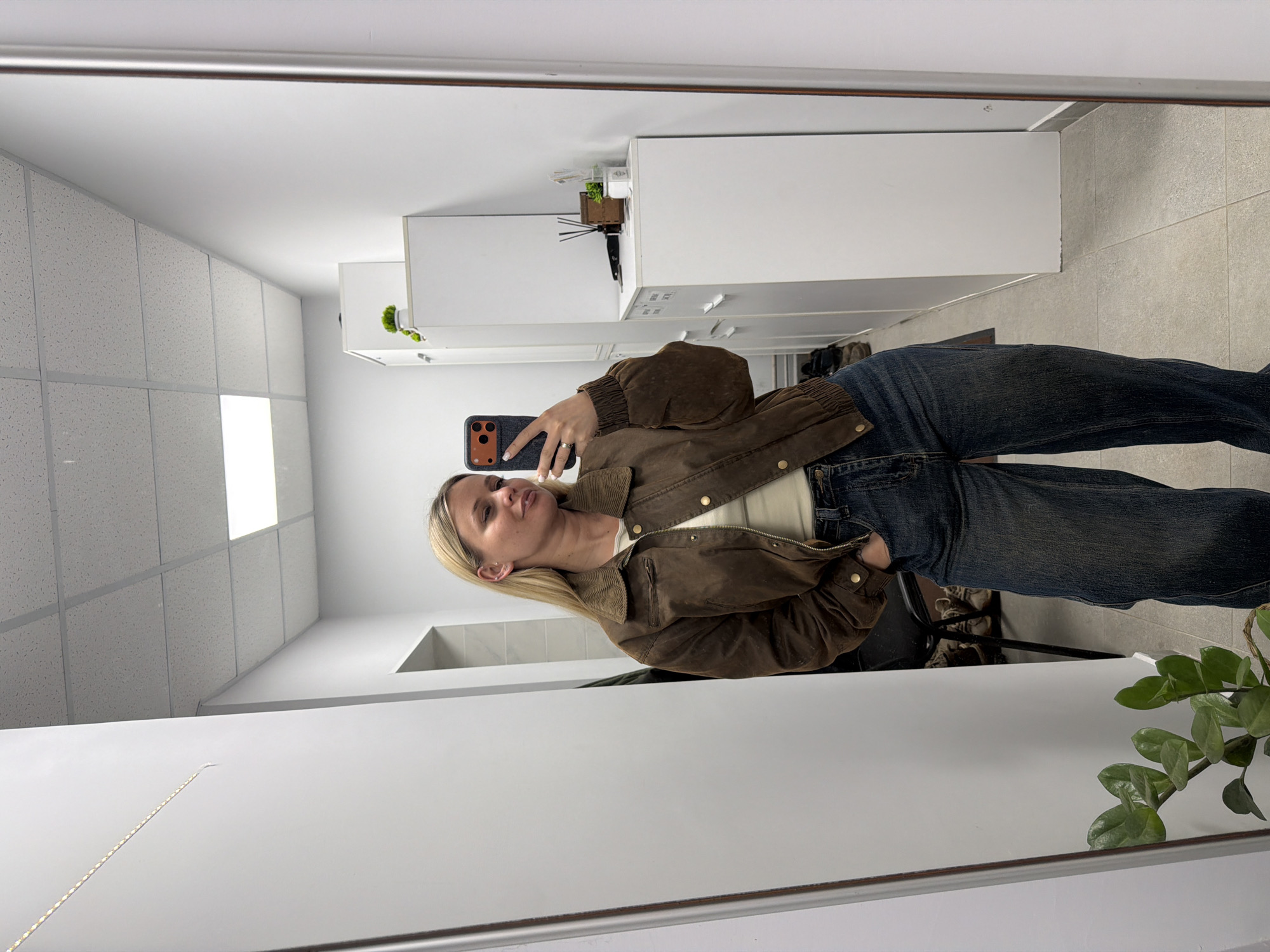 A blonde woman is taking a selfie in a mirror. She is wearing a brown jacket, a white shirt, and blue jeans. The background includes a white countertop and cabinets, a tiled floor, and a houseplant. The location is not identifiable as Sarapul, Russia.