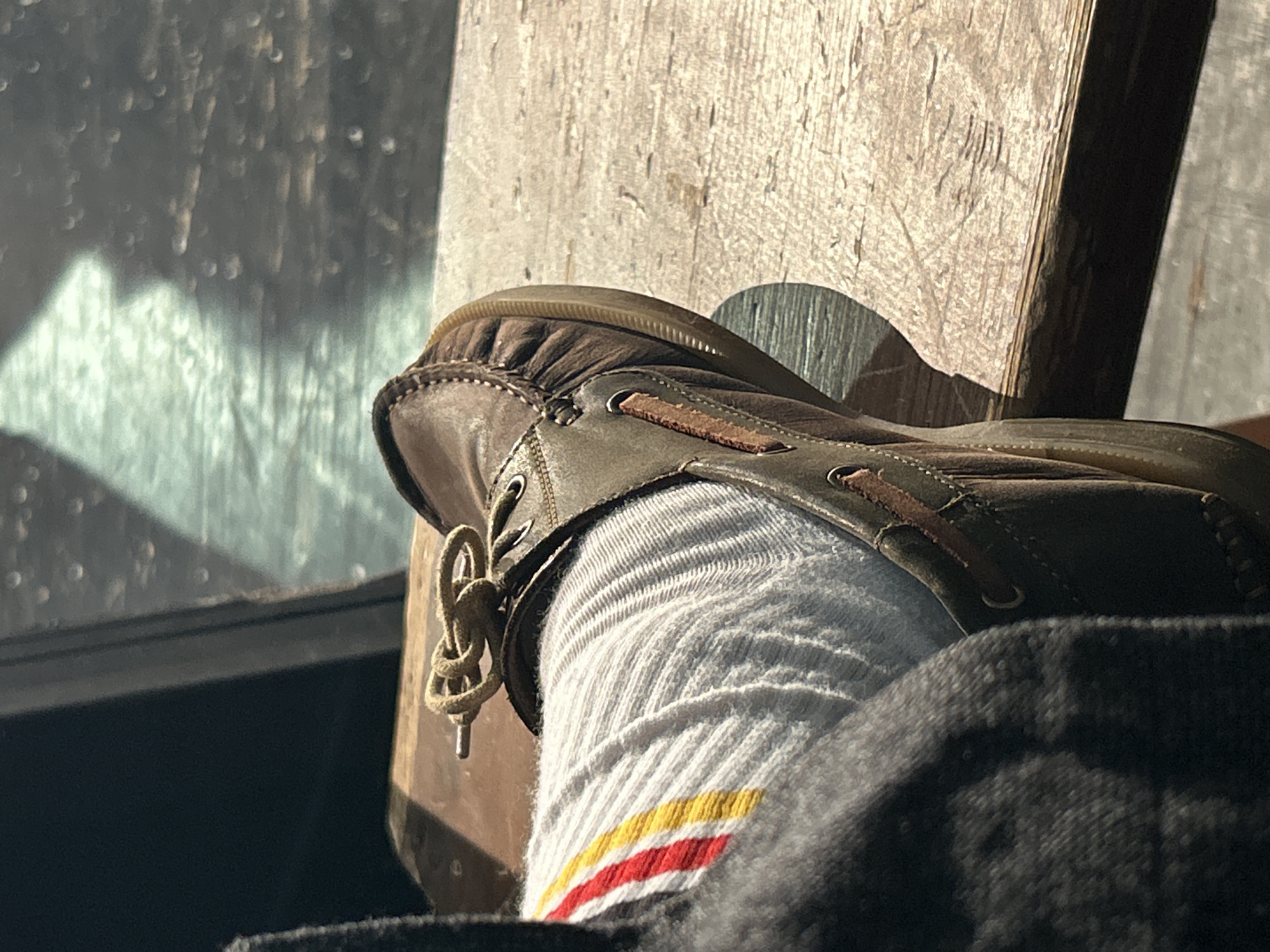 A close-up shot focuses on a person's foot and lower leg, clad in a brown leather boat shoe and a ribbed white sock with red and yellow stripes. The shoe is tied with a tan lace. The foot rests on a weathered wooden surface, likely a bench or chair, which is illuminated by bright sunlight, casting a distinct shadow. To the left of the shoe, a window with water droplets on its surface is visible, creating a blurred, bokeh effect. The light filtering through the window suggests it is daytime, possibly morning or afternoon, and the presence of water droplets hints at recent rain or condensation. The overall mood is relaxed and casual, with the warm lighting adding to the cozy atmosphere. There is no visible text in the image.