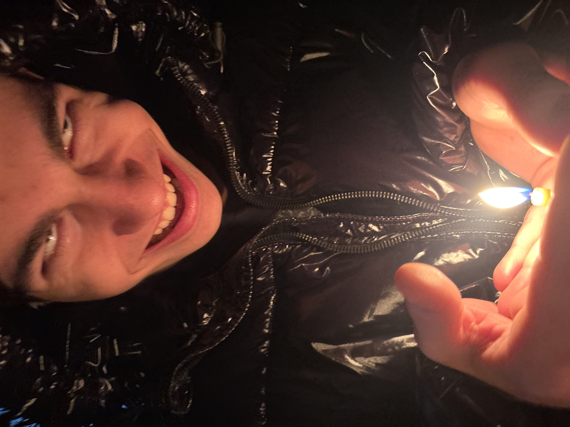 A person is angled, smiling widely, wearing a shiny black puffer jacket with the zipper partially visible. The person is holding a lit lighter in their hand, with the flame visible. The background is dark and appears to be indoors. There is no information about the city of Lehchevo, Bulgaria in the image.
