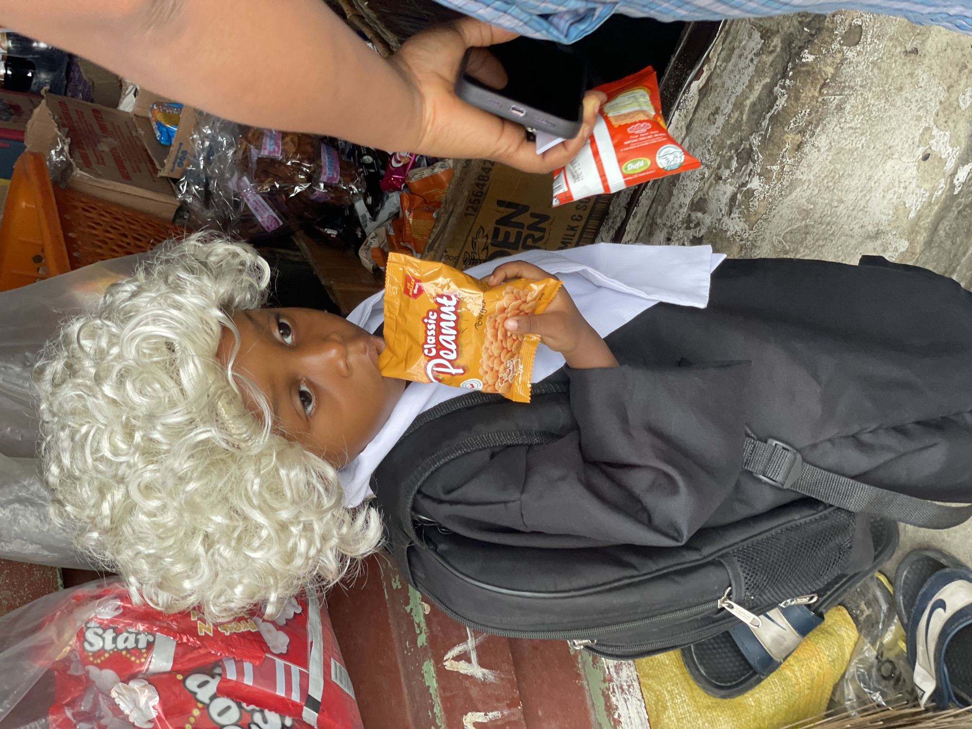This image captures a candid moment in what appears to be a bustling outdoor or semi-outdoor setting, likely a market or cluttered shop area in Ado, Nigeria. The central subject is a young child, possibly of preschool or early primary school age, with dark skin, who is dressed in a distinctive costume. The child wears a large, curly white wig, resembling a judge's or barrister's wig, over their head, paired with a white collar and a black robe-like garment. They are lying down, looking towards the viewer, and are actively eating "Classic Peanut" snacks from an orange packet.
Above the child, an adult's arm and hand are visible, holding a smartphone, suggesting they are taking a picture or video of the child. The adult also holds a small red snack packet, possibly labeled "Dufil." Next to the child is a black backpack, and nearby, a pair of blue and white Nike slides can be seen.
The background is dense with various items, indicating a commercial environment. Visible objects include clear plastic bags filled with snacks, red snack packets labeled "Star" and "Auto N," and cardboard boxes with partially discernible text such as "12564842," "OPEN RN," and "MILK & SO." The ground beneath is uneven and worn, with peeling paint in patches of red, yellow, and green, consistent with a well-used public or commercial space. The image portrays a casual, daytime scene, possibly during a school event, dress-up play, or a family outing, with the child enjoying a snack.