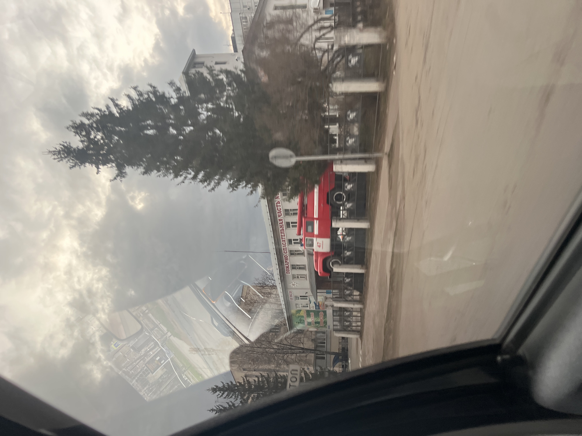 The image presents an outdoor urban scene in Chaikovsky, Russia, viewed from inside a vehicle, likely through a side window or sunroof. The primary subject is a multi-story building, appearing to be a fire station. A large red fire truck is prominently parked in front of this building.
On the building's facade, large Cyrillic text reads "ПОЖАРНО-СПАСАТЕЛЬНАЯ ЧАСТЬ №", which translates to "Fire and Rescue Unit No.". Below this, other partially visible signs include words like "СПАСЕНИЕ" (Rescue) and "ПОМОЩЬ" (Help), reinforcing the building's purpose. The red fire truck has the number "47" clearly visible on its side.
The sky is heavily overcast with dense clouds, suggesting a cloudy daytime setting. To the left of the fire station, a large tree, possibly evergreen or winter-bare with dark branches, stands tall. The ground in the foreground appears to be a road or paved area. The bottom right and left of the image show parts of the vehicle's dark interior frame and glass. Significant reflections on the glass surface create a superimposed layer, showing what resembles an inverted aerial map or cityscape over the actual scene. The overall impression is a mundane yet functional streetscape in a Russian town.