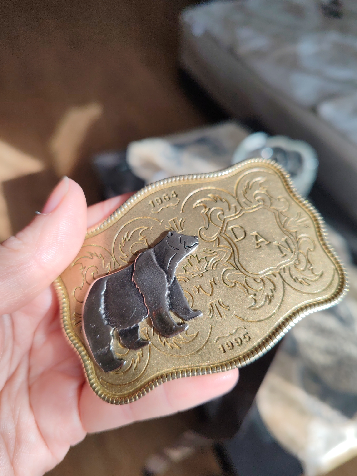 A person's hand, positioned on the left side of the frame, holds a decorative metal belt buckle, likely indoors in Saint Petersburg, Russia. The buckle features a prominent gold-toned base with an ornate, scalloped edge. A dark, silver-colored bear figure is overlaid on the left side of the buckle, depicted in a standing posture, facing left.
The gold surface of the buckle is extensively engraved with intricate swirling patterns and small, possibly maple leaf-like designs. Two distinct years are inscribed on the buckle: "1964" is visible at the top left corner, and "1995" appears at the bottom right. Centered on the right side of the buckle, within a shield-like emblem adorned with additional engravings, are the block letters "DAN". The buckle's perimeter features a textured, ridged, or beaded border. The background of the image is out of focus, indicating an indoor setting with diffused lighting that casts reflections on the metallic surfaces of the buckle.